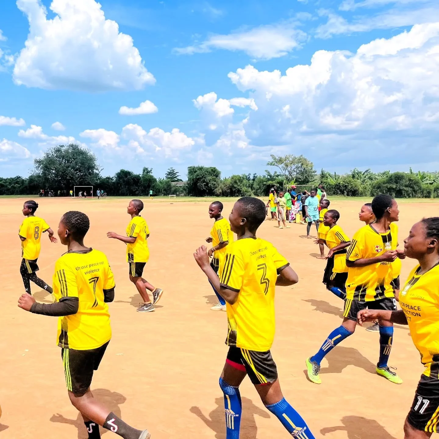 Children in yellow soccer jerseys running on a dirt field during daytime with blue sky and clouds, and trees in the background.