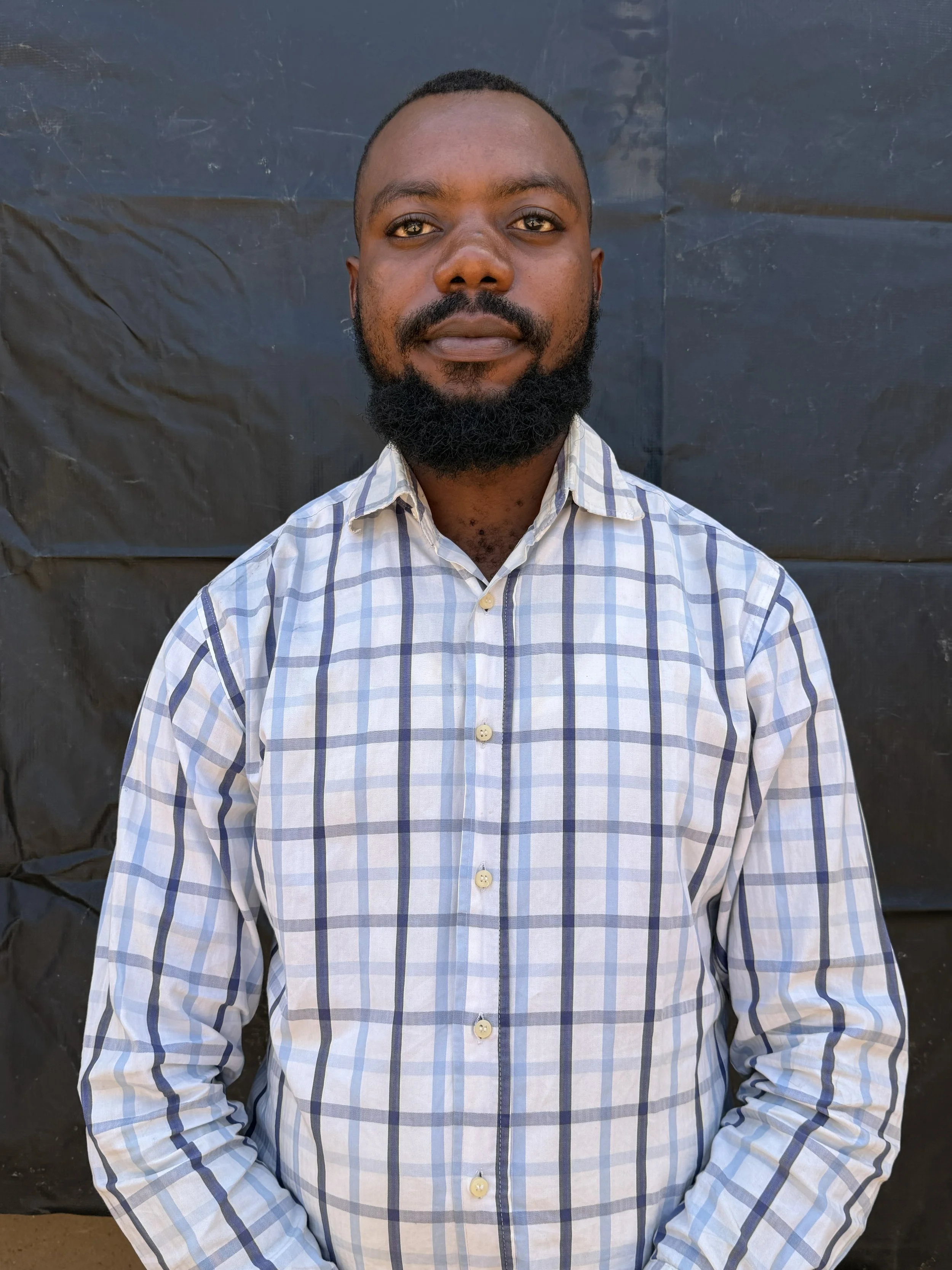 A portrait of a man with dark skin, a short beard, and short hair, wearing a plaid shirt, standing in front of a black background.