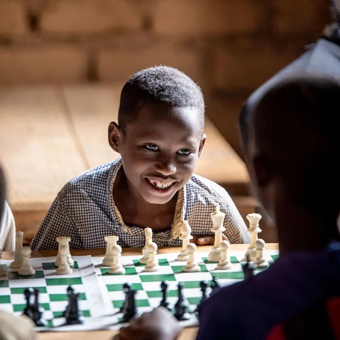 Young boy with a broad smile playing chess with another person at a wooden table in a rustic room.