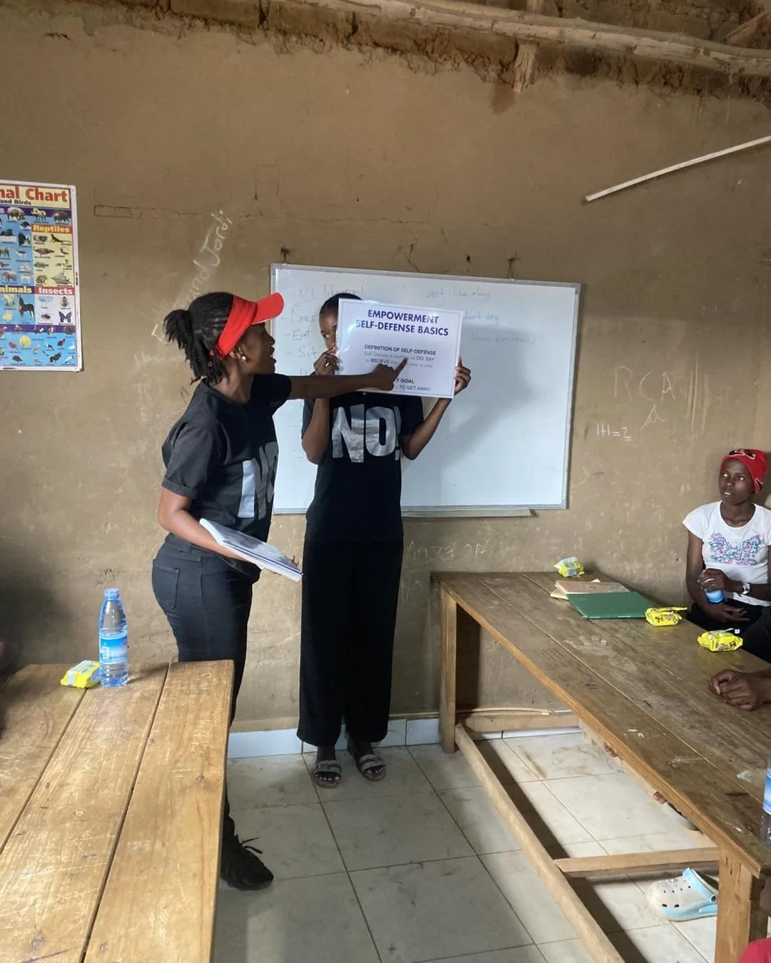 Two women standing in front of a whiteboard in a classroom, one is holding a sign about self-defense basics, and others are sitting at wooden tables with snacks and water bottles.