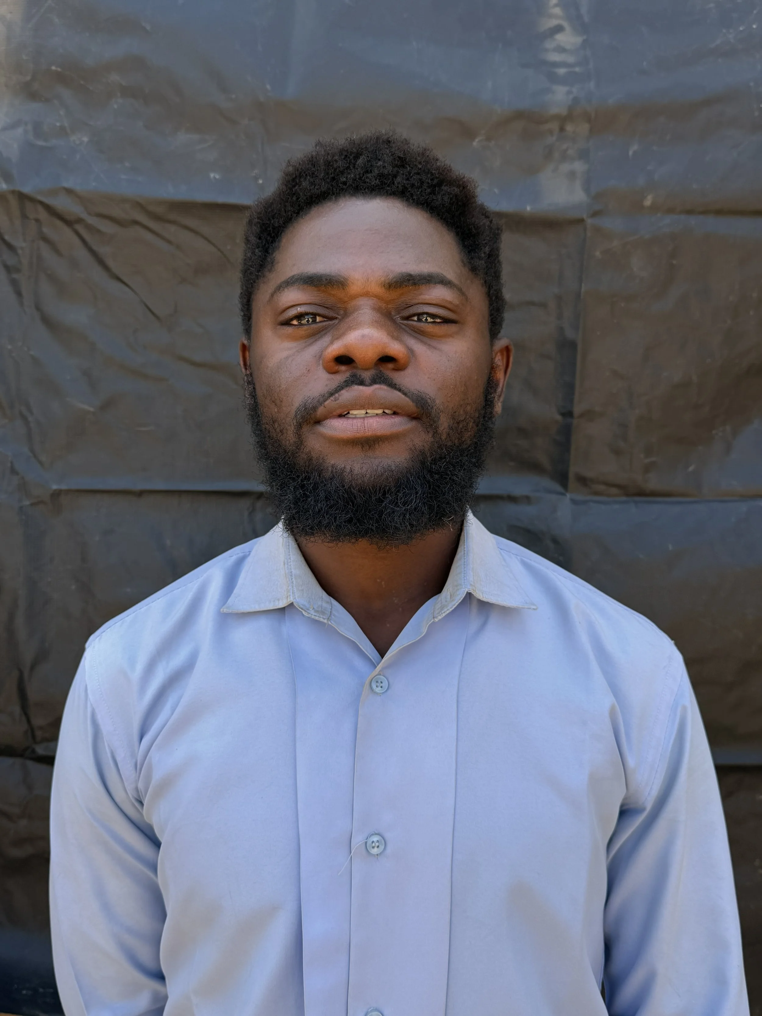 Close-up of a young man with dark skin, curly black hair, and a full beard, wearing a light blue button-up shirt, standing in front of a textured dark background.