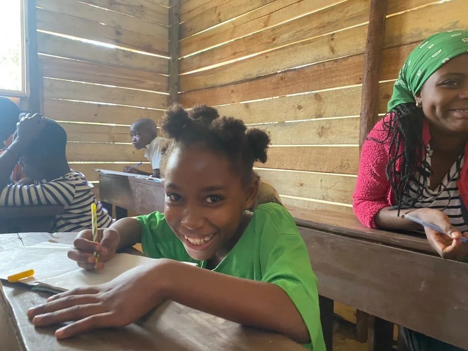 A young girl with a bright smile, sitting at a wooden desk in a classroom, holding a yellow pencil. Other children are visible in the background, sitting at similar desks inside a wooden-paneled classroom.