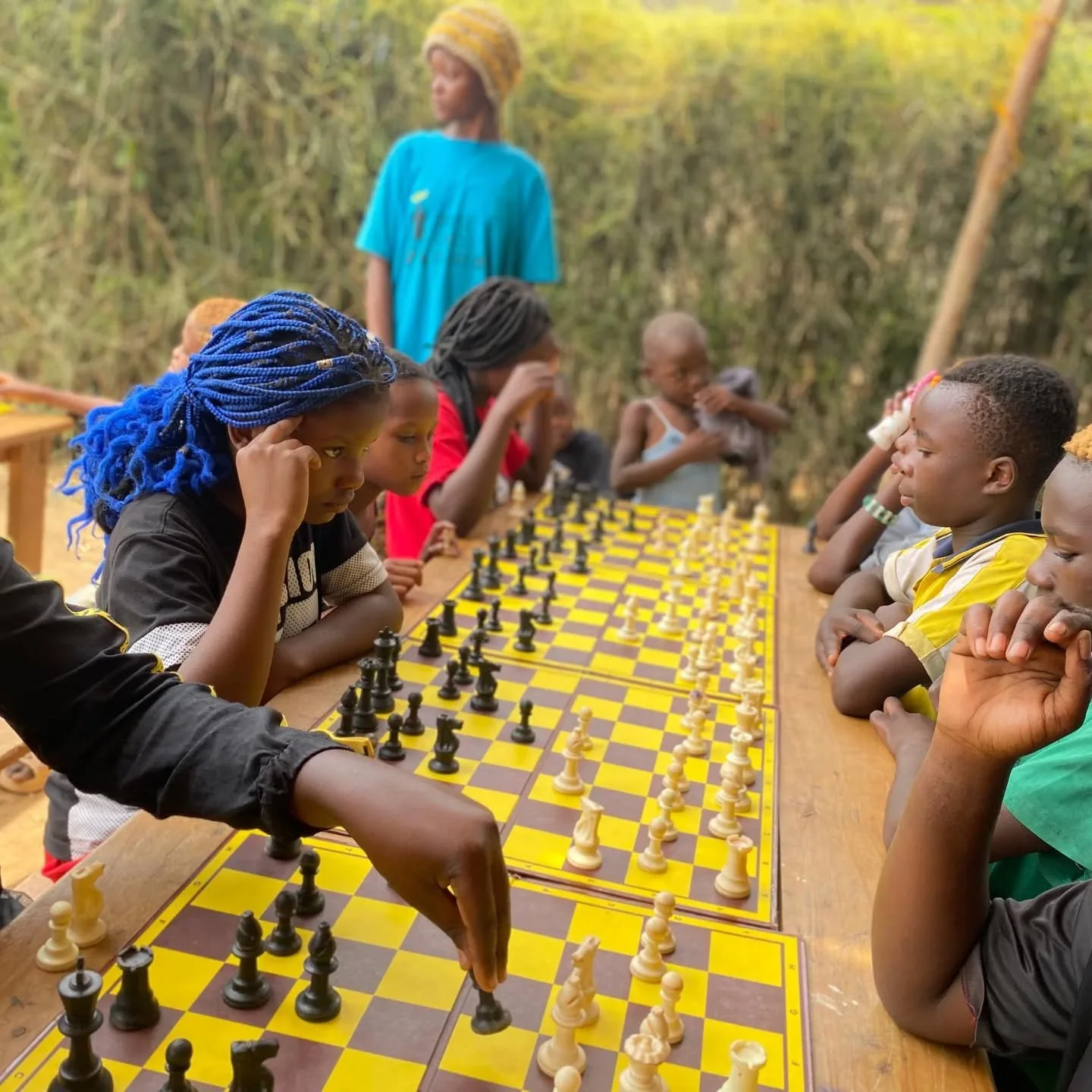 Children sitting on each side of a chessboard outdoors, playing chess while several children observe, with trees in the background.