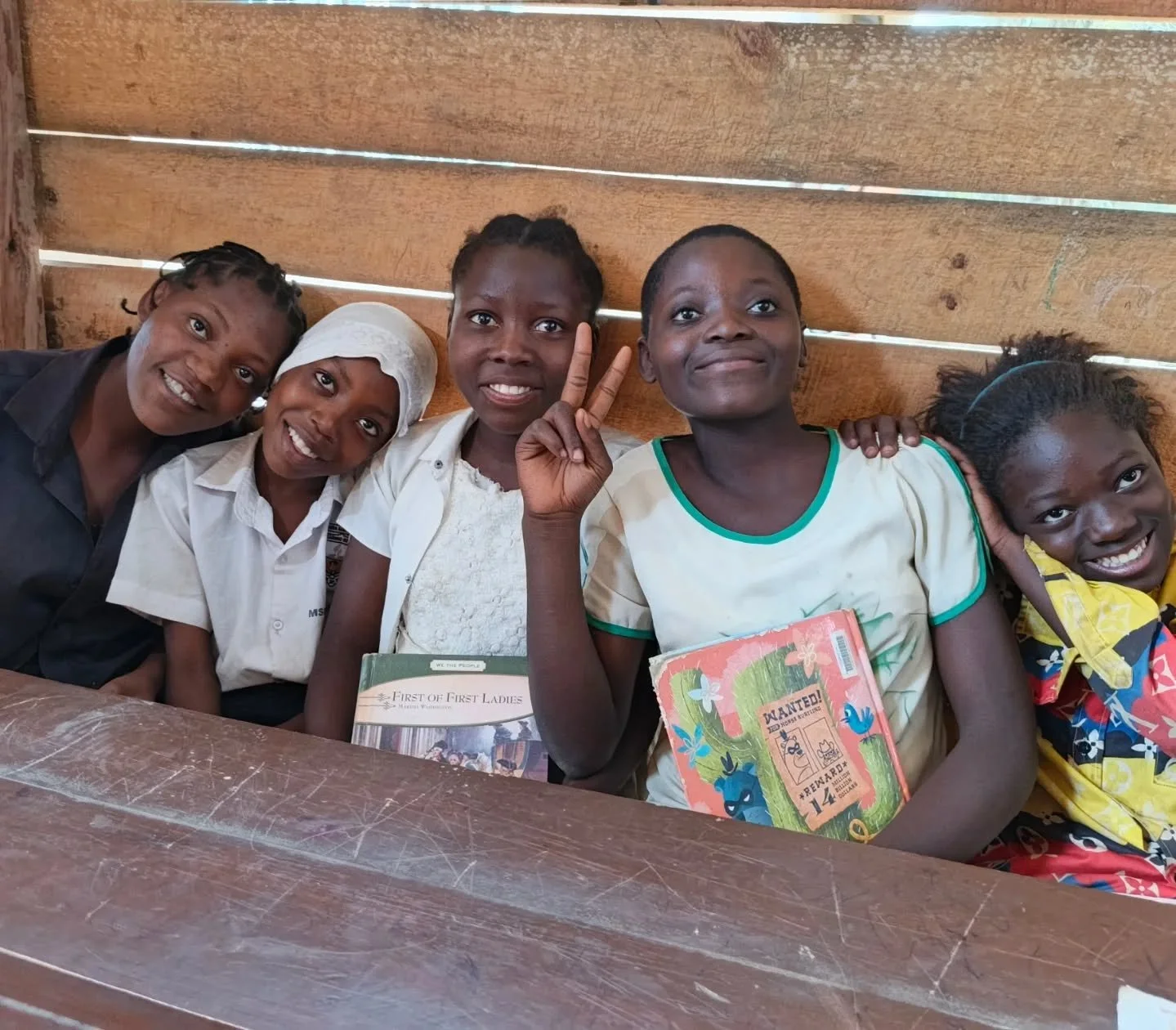 Five smiling young girls sitting together in a wooden classroom. One girl in the middle is making a peace sign with her hand and holding a colorful book. The background is wooden and the girls are sitting behind a wooden desk.