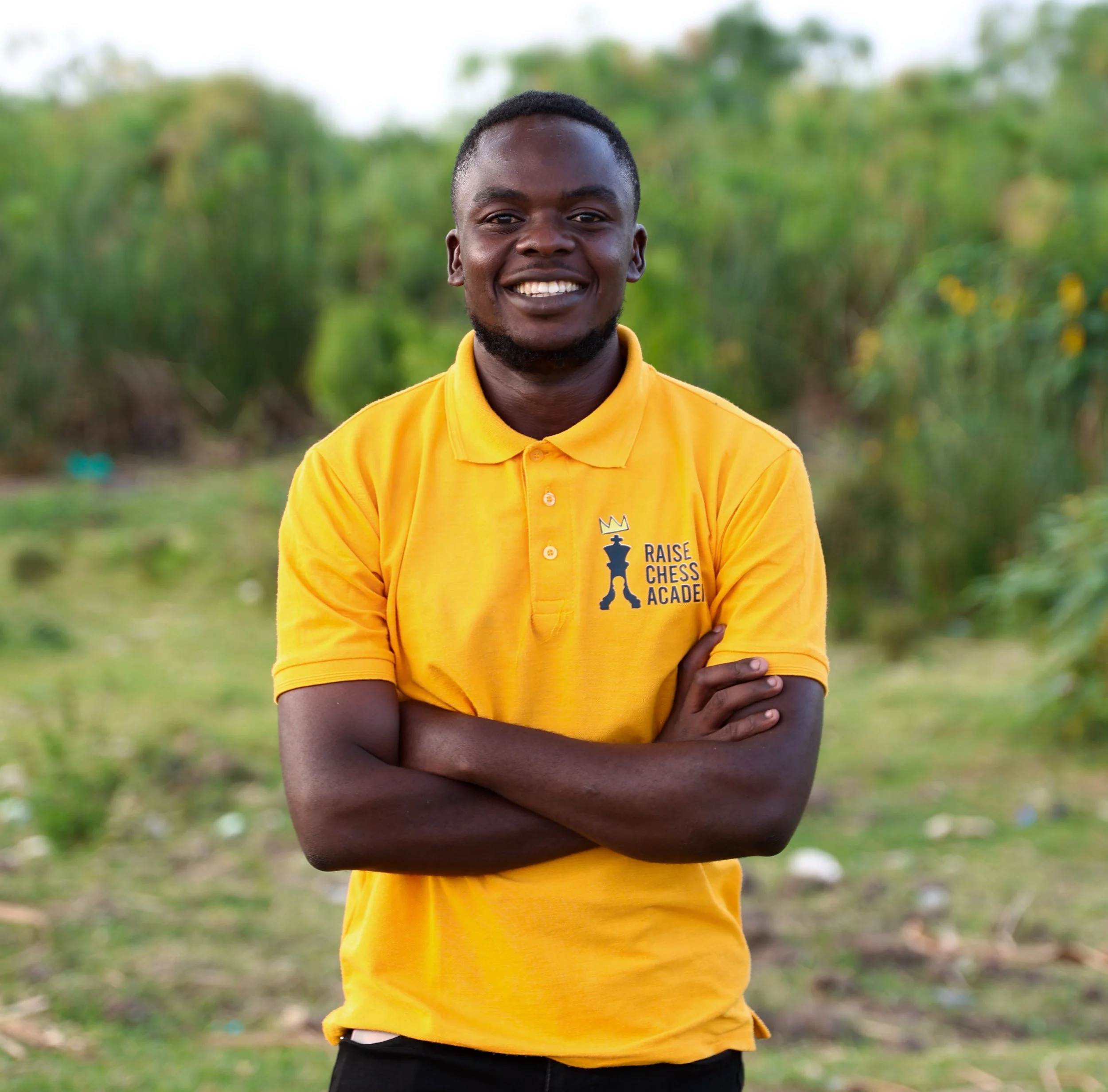 Young man with dark skin, short hair, and beard smiling, wearing a yellow polo shirt with 'Raise Chess Academy' logo, standing outdoors with green foliage background.
