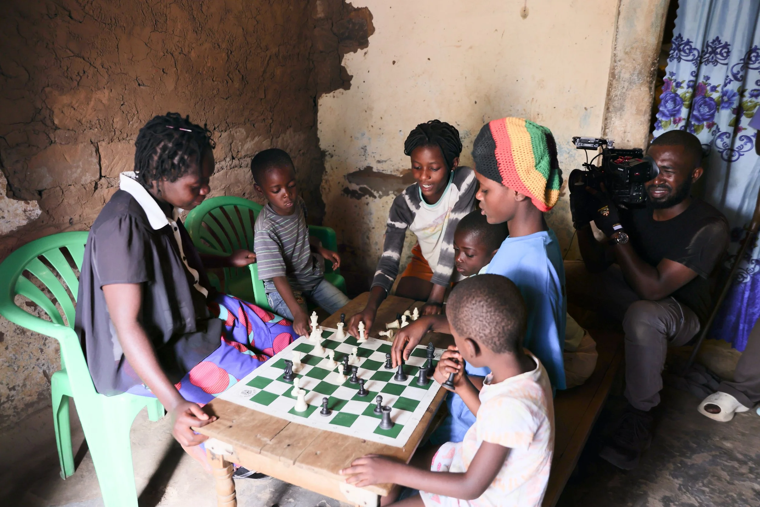 Children and adults playing chess around a wooden table in a modest room, with one person filming the game, and colorful curtains hanging beside a textured wall.