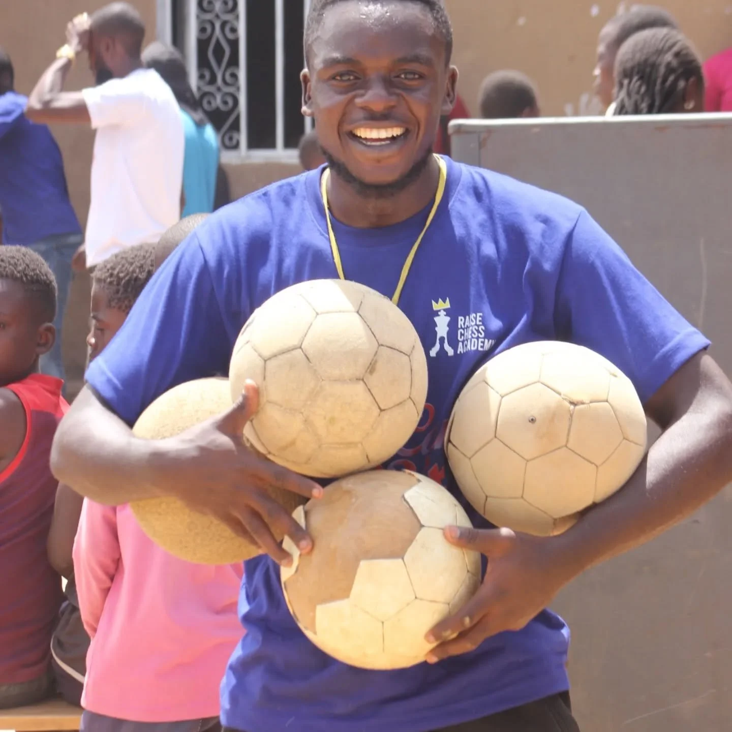 Man smiling, wearing a blue shirt with 'Raise Chess Academy' logo, holding several old, yellowed soccer balls, outdoors with children and people in background.