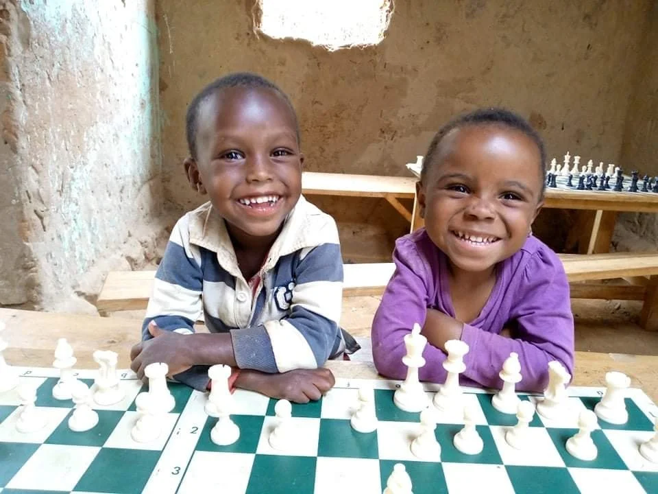 Two smiling children sitting at a table playing chess with a rustic wall and additional chess set in the background.