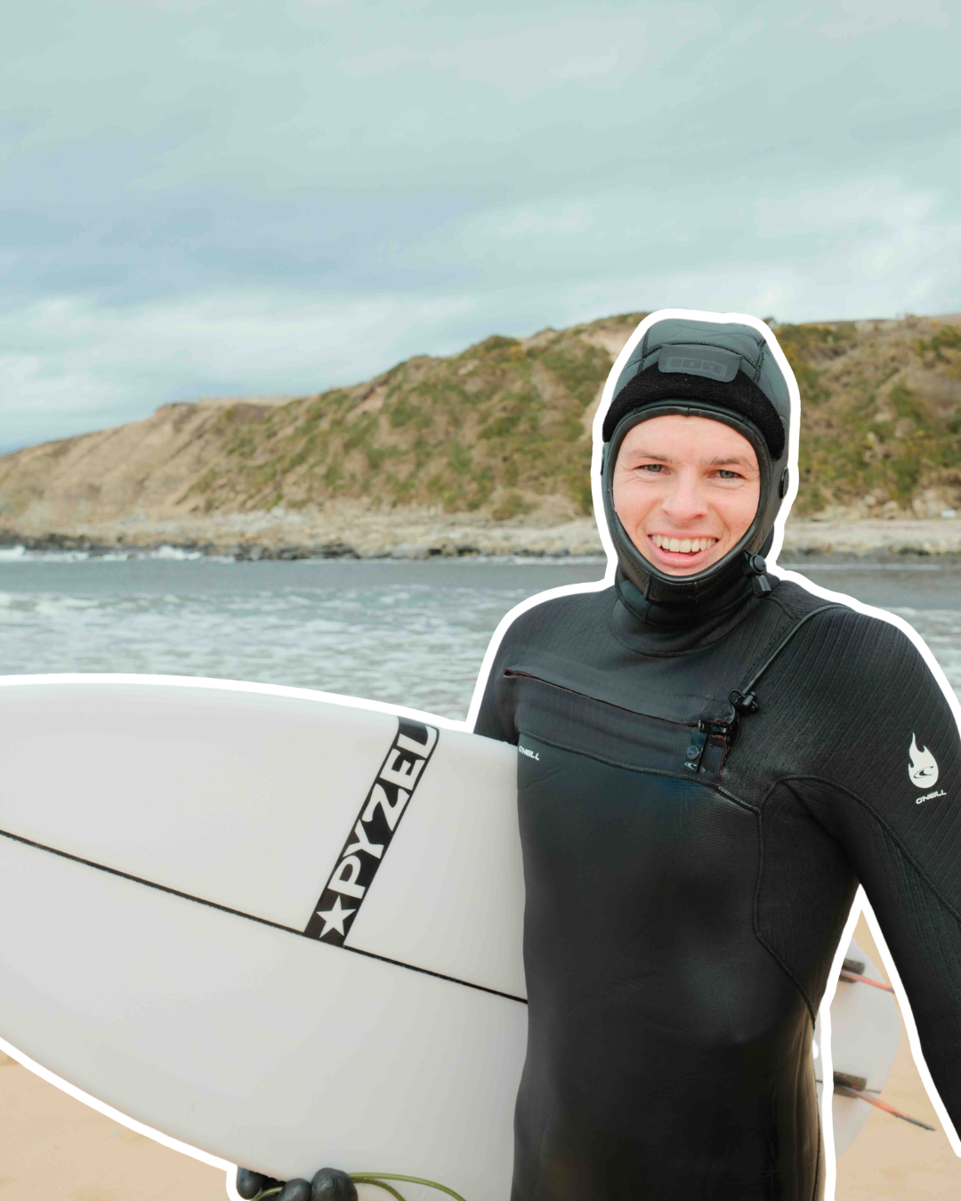 A man in a black wetsuit holding a white surfboard on a beach with rocky cliffs and cloudy sky in the background.