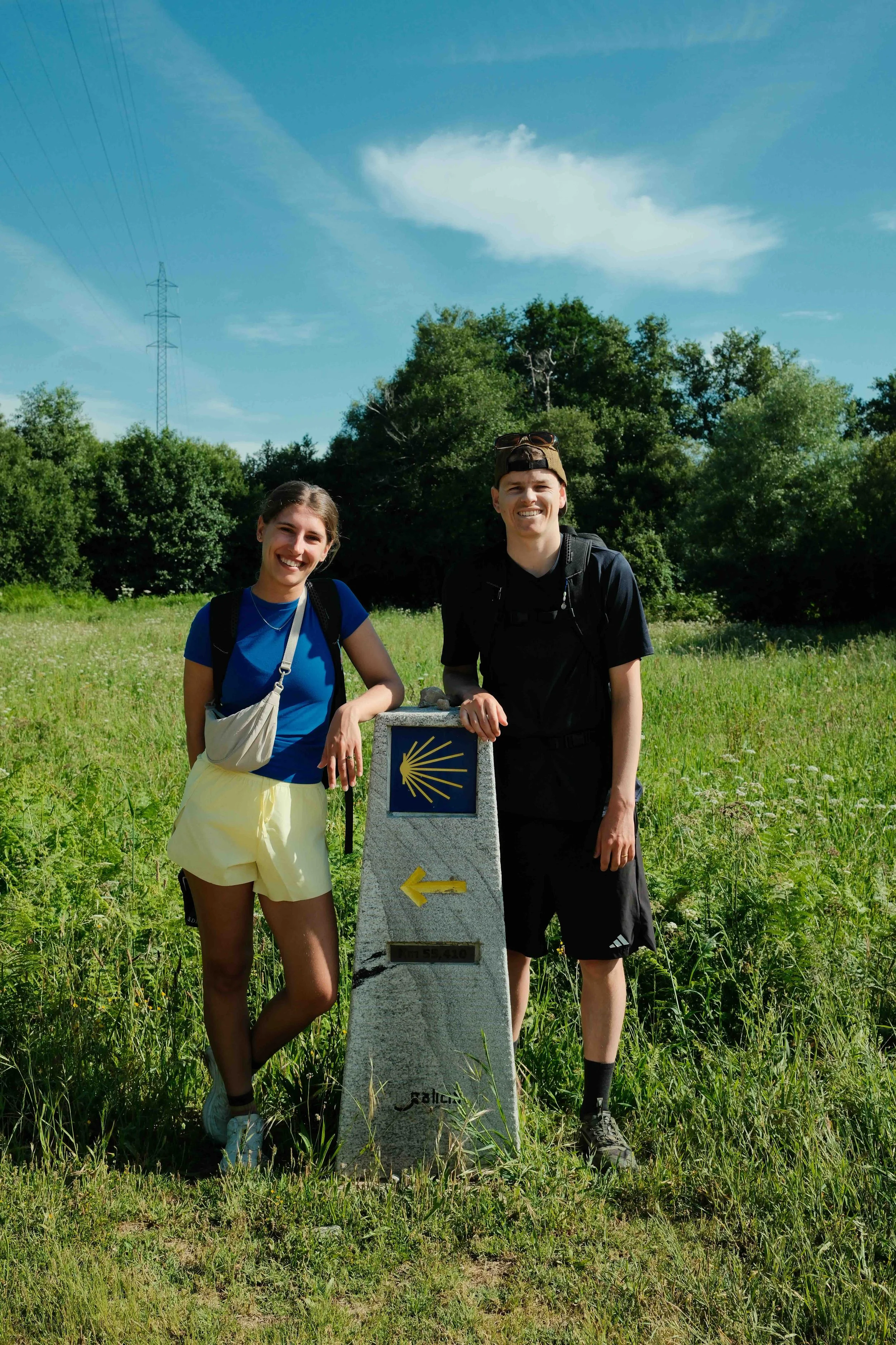 Two young people, a woman and a man, standing outdoors on a grassy field beside a trail marker with the Shell logo and an arrow pointing left, smiling at the camera. The woman is on the left, wearing a blue top, yellow shorts, and has a white bag across her shoulder. The man is on the right, wearing a black shirt and black shorts, with sunglasses on his cap. In the background, there are green trees, a blue sky with some clouds, and an electrical tower.