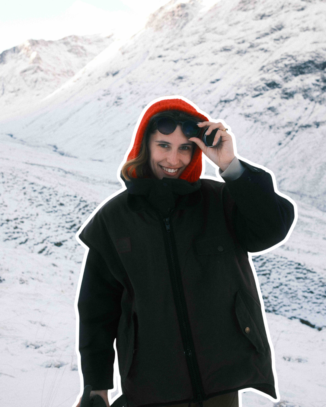 A woman in winter clothing, including a black jacket and red knit hat, outdoors in a snowy mountainous landscape, smiling while holding sunglasses.