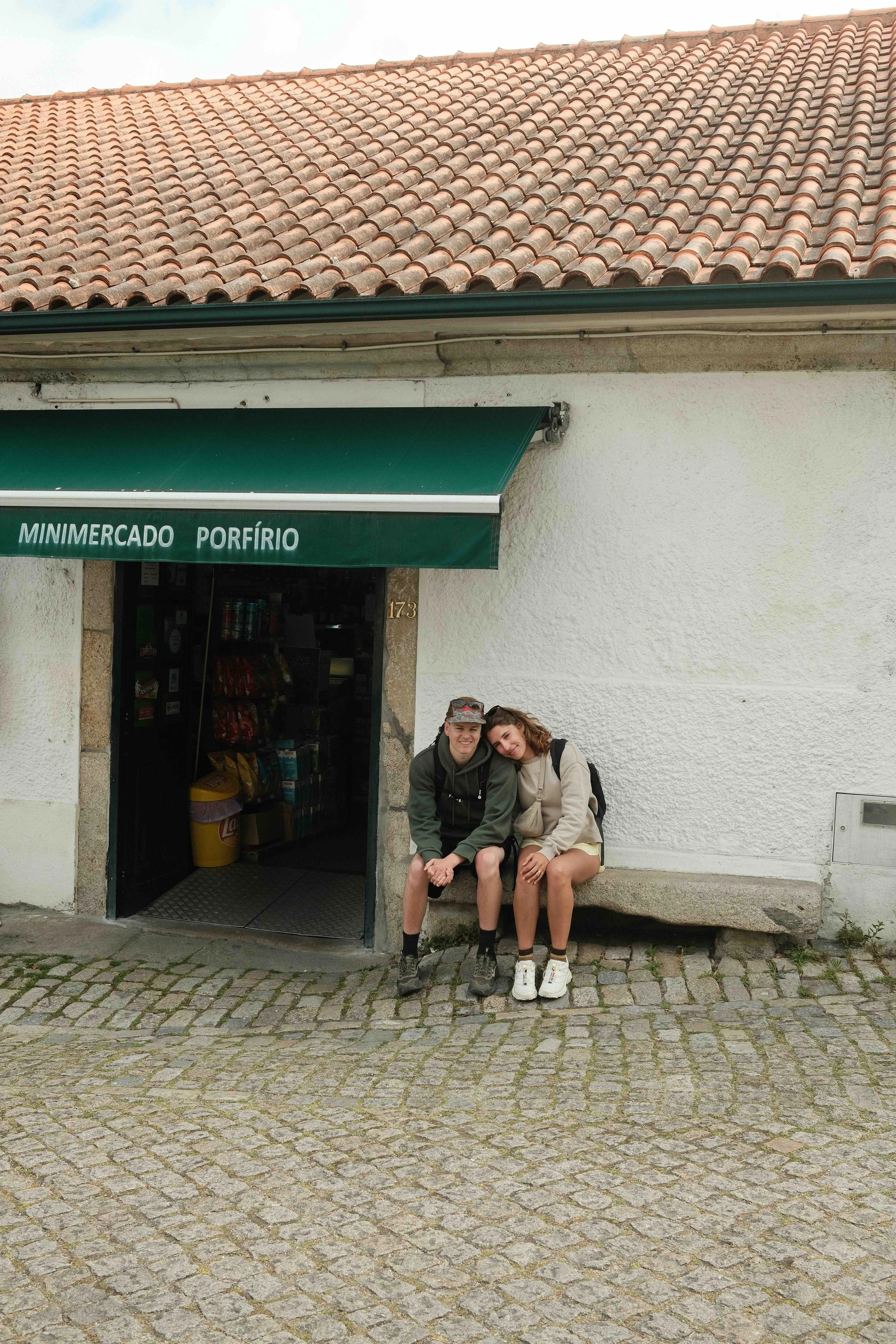 Two young people sitting on a stone bench outside a small grocery store called 'Minimercado Porfírio.' They are close together, smiling, with a building with a tiled roof in the background and a cobblestone street in the foreground.