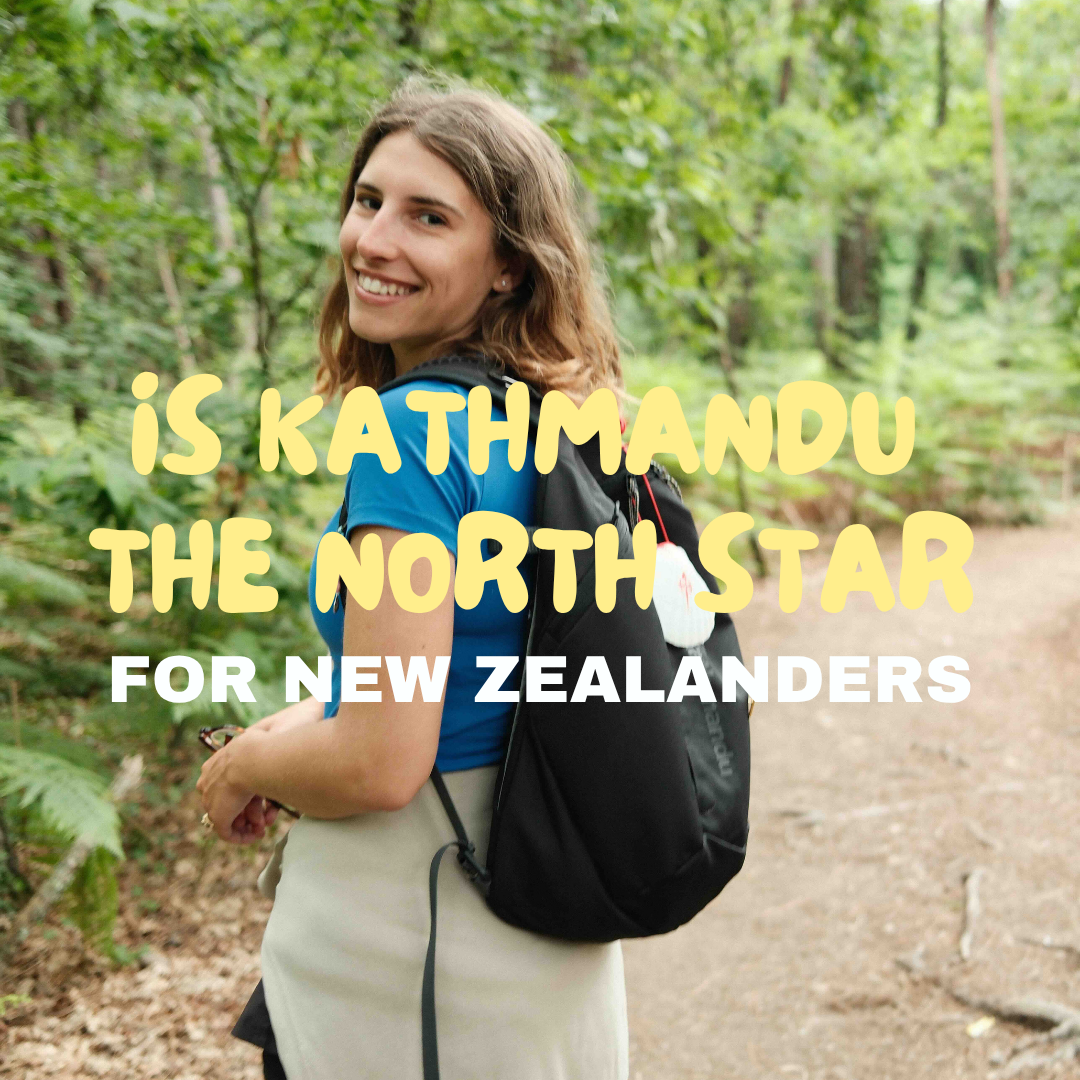 A smiling woman with brown hair and a blue shirt standing on a forest trail with trees and greenery in the background, carrying a black backpack.