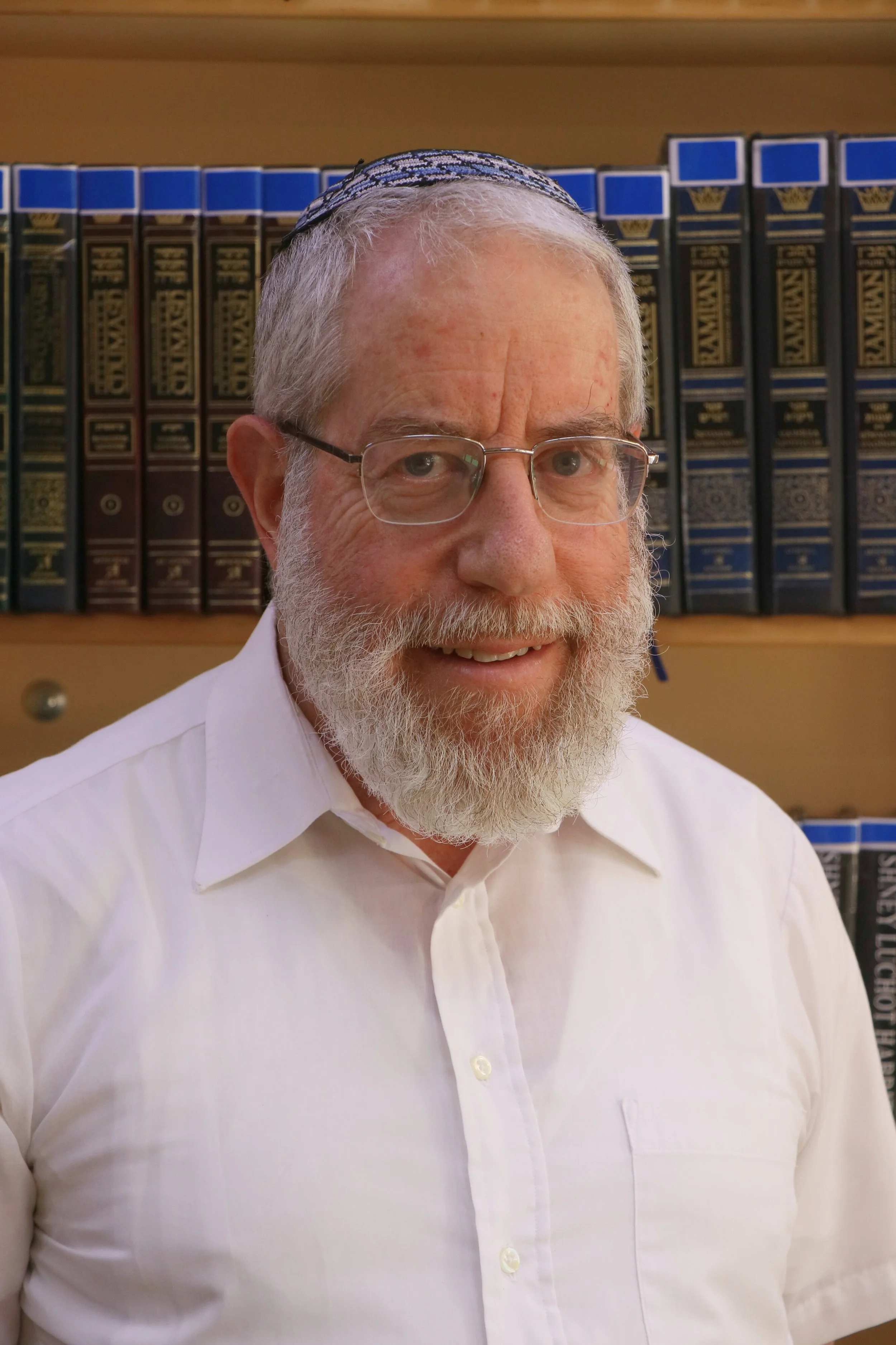 Close-up of an elderly man with glasses, a white beard, wearing a white shirt and a blue kippah, standing in front of a bookshelf filled with books.