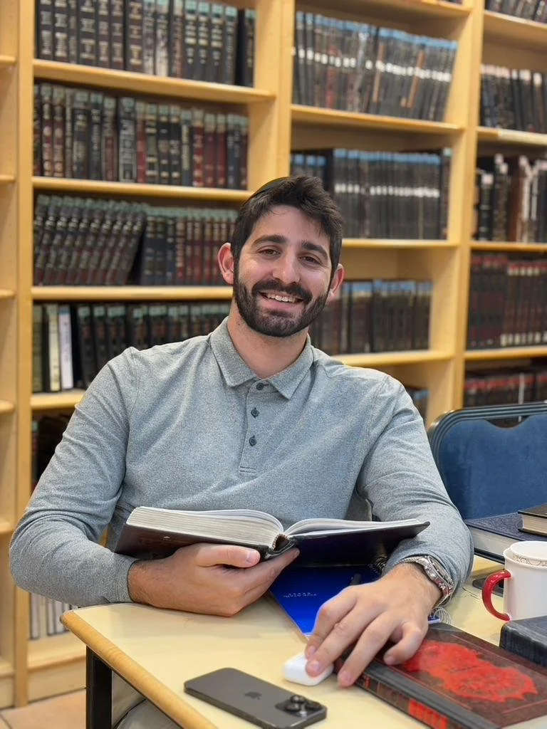 A smiling man with dark hair and a beard, wearing a gray collared shirt, sits at a table in a library. He has an open book in his hands, with shelves of books in the background.