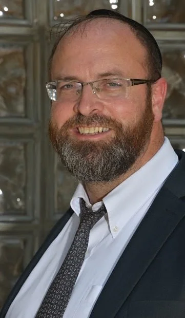 A man with glasses, a beard, and short hair wearing a suit, white shirt, and patterned tie, smiling at the camera.
