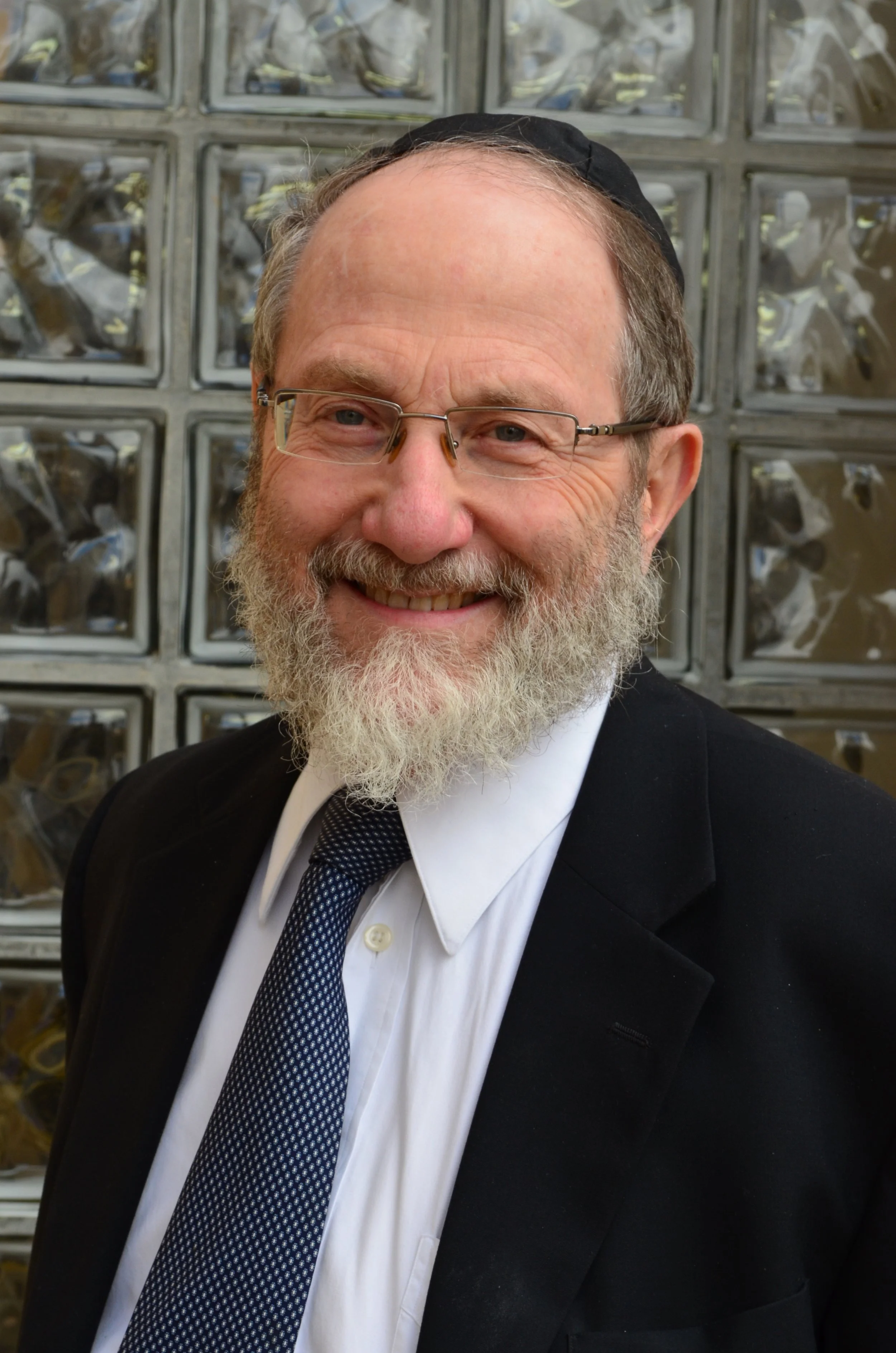 A man in a black suit, white shirt, and dark patterned tie smiling, with glasses, beard, and a black skullcap, in front of a glass block wall.