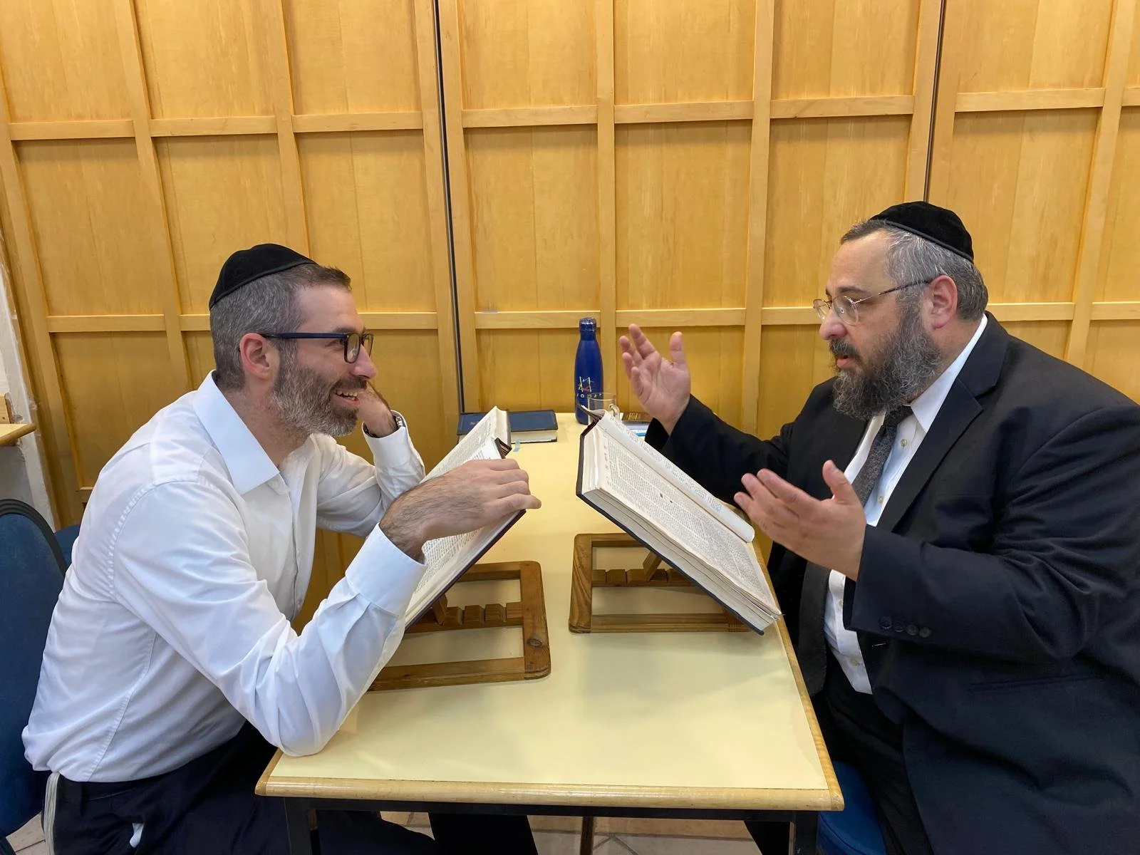 Two men with beards and glasses, wearing yarmulkes, sitting at a table, engaged in a discussion and reading from large open books, with notebooks and a water bottle on the table, in front of a wooden-paneled wall.