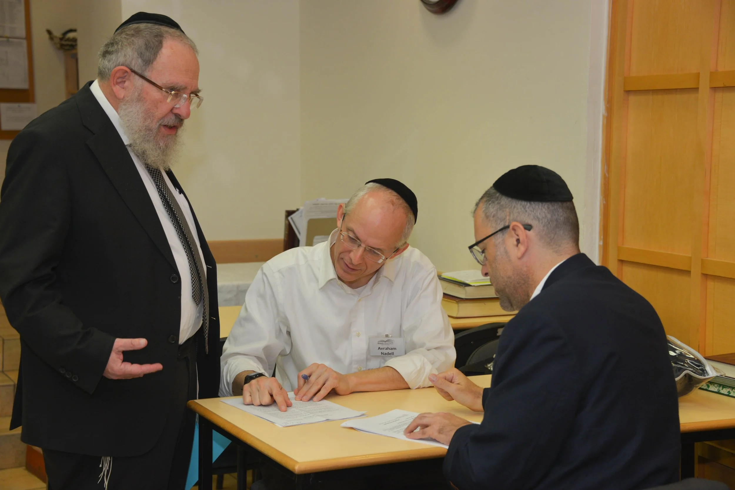 Four men, three seated and one standing, are engaged in a discussion over documents at a table in a room with beige walls and wooden furniture. Two of the men are wearing yarmulkes.