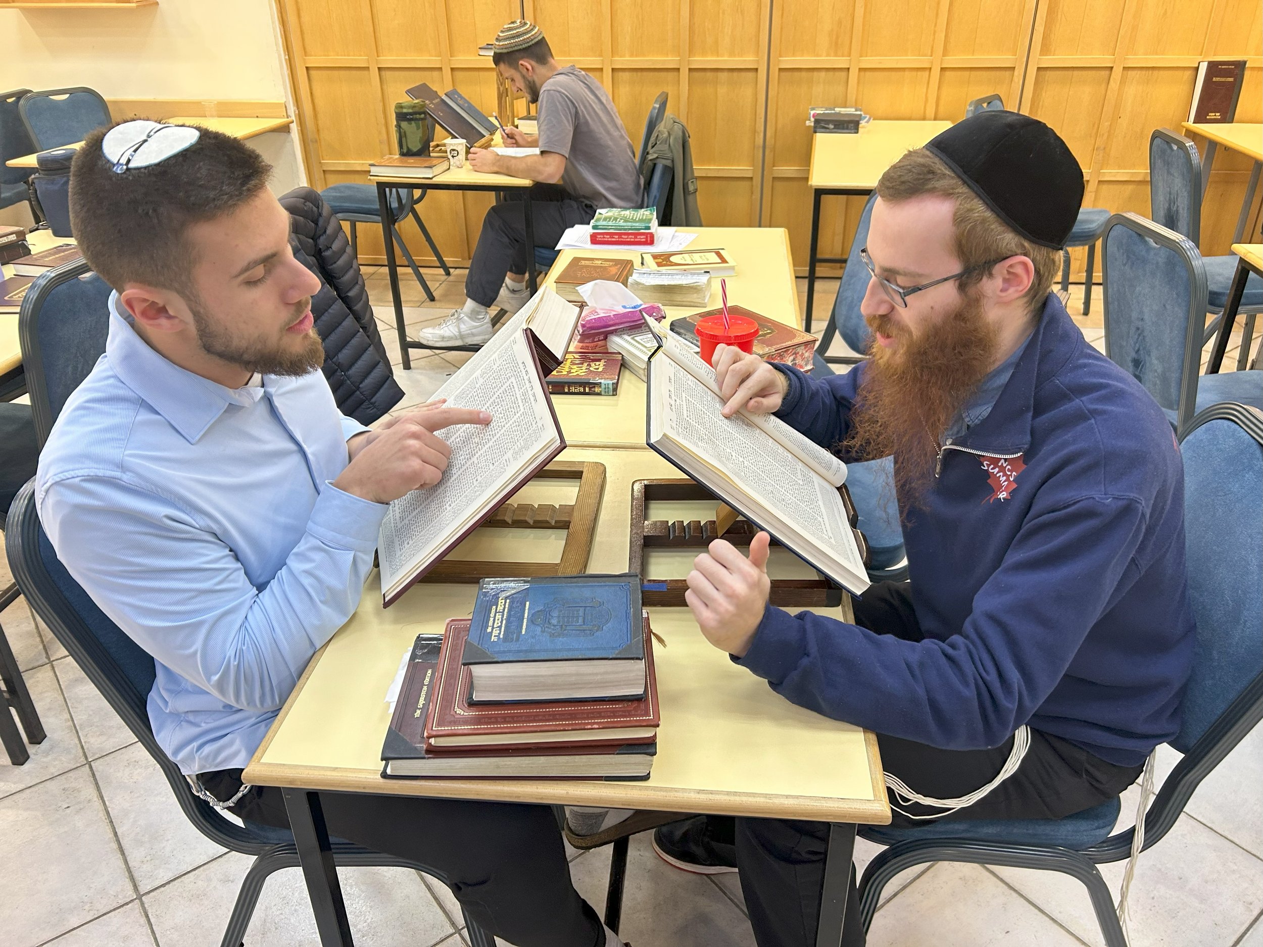 Two young men sit across from each other at a table, reading from large books, with more books piled on the table. A third person in the background is sitting at a separate table, also reading, with books and a cup on the table. All are inside a room with wood-paneled walls and blue chairs.