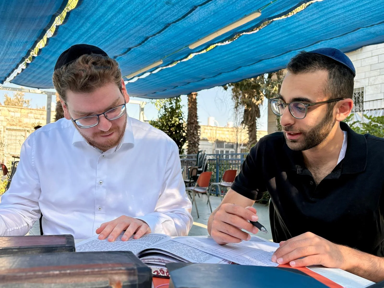 Two men, wearing kippahs and glasses, are sitting outdoors at a table looking at a book or document together. There are chairs and trees in the background.
