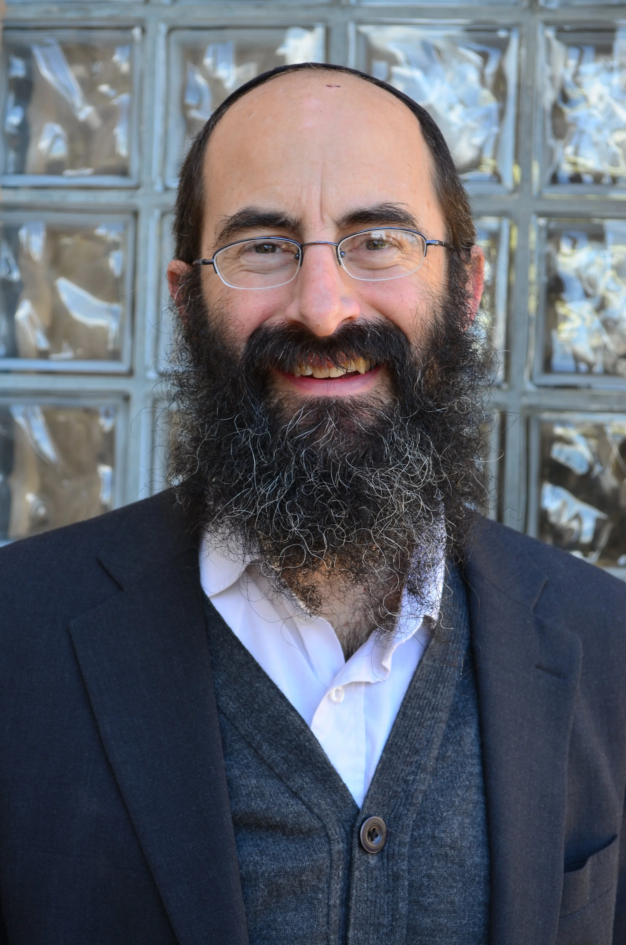 Close-up of a smiling man with glasses, a full beard, wearing a dark suit and a white shirt in front of a modern glass block wall.