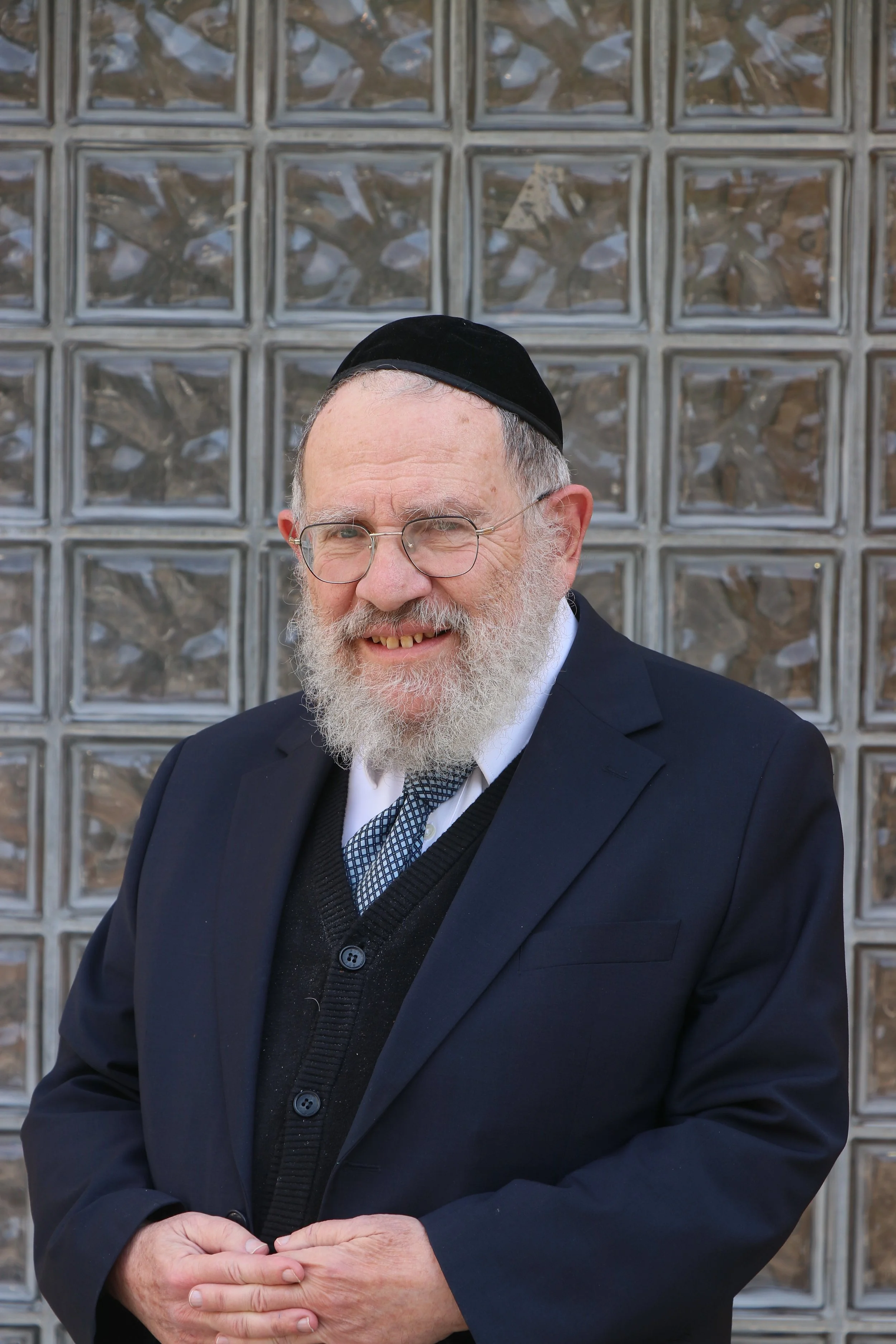 An elderly man with glasses and a white beard, wearing a black kippah and a dark suit, standing in front of a glass block wall.