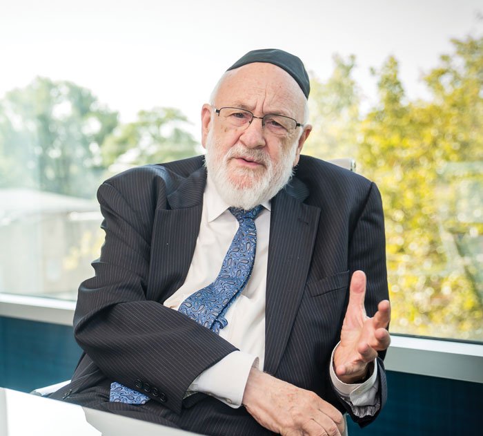 An older man with a white beard and glasses, dressed in a suit and tie, sitting in an office with large windows and trees outside, wearing a kippah.