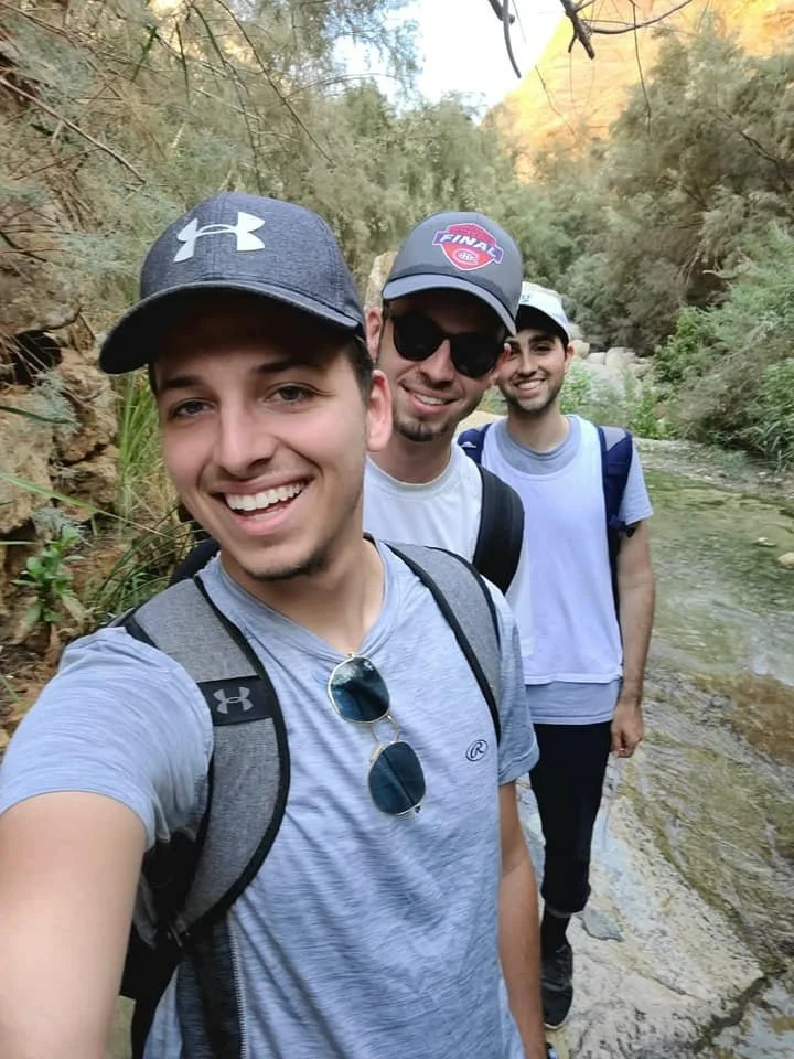 Three young men are taking a selfie together outdoors near a stream, with trees and hills in the background, during a hike in a natural setting.