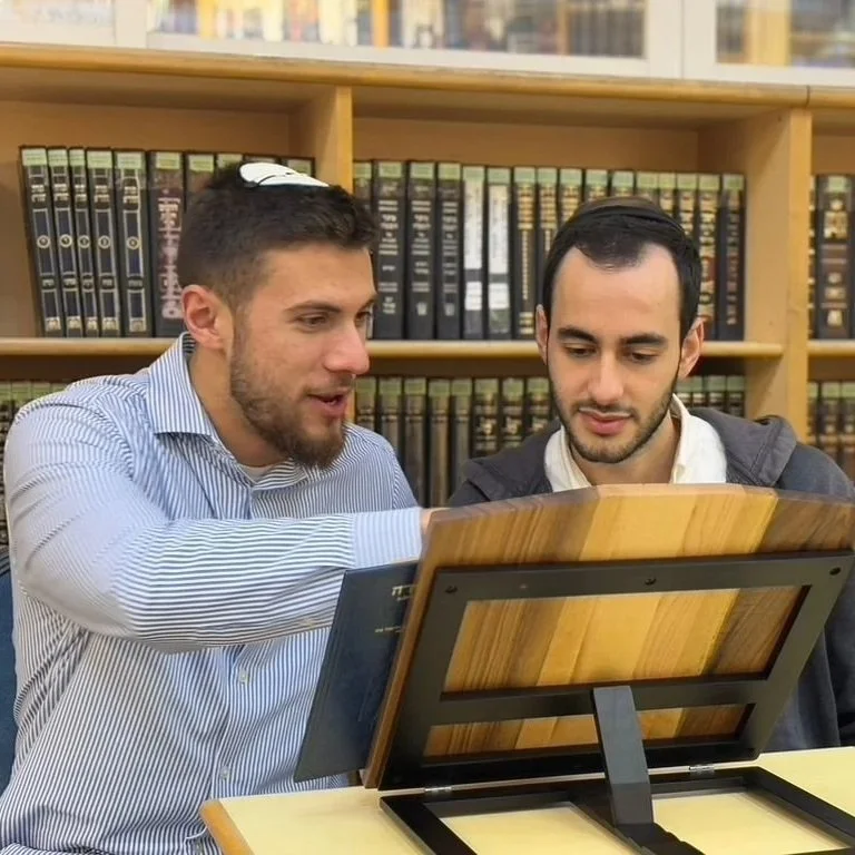 Two men in a library looking at a book or a document on a wooden table, with bookshelves filled with books behind them.