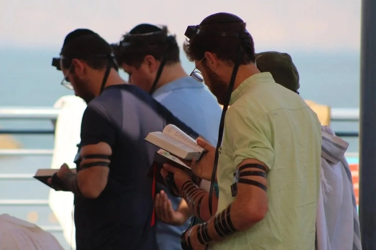 Four people stand close together reading books inside a shaded area near a body of water, with a railing behind them.