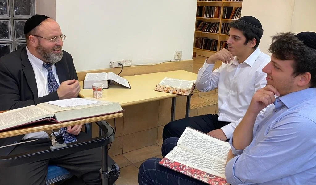 A man with a beard, glasses, and a black kippah is speaking and gesturing, seated at a table with open books and a paper cup. Two young men wearing dark kippahs, white and light blue shirts, are listening attentively, each with an open book on their laps, in a room with a bookshelf in the background.