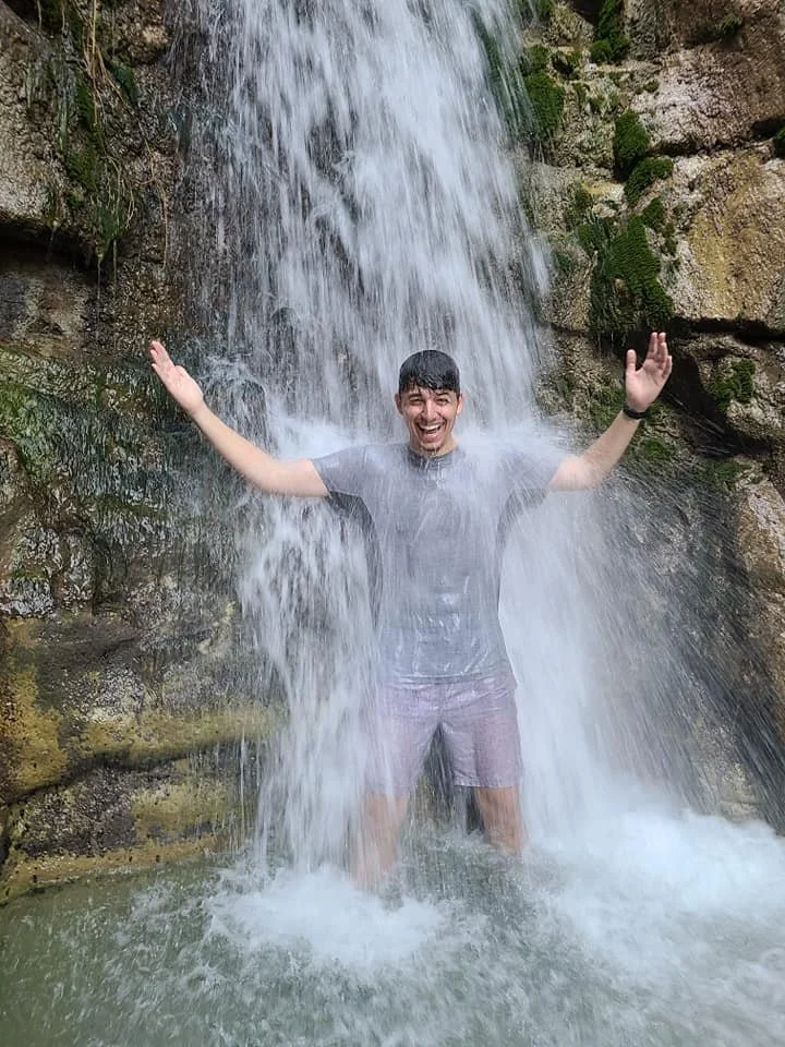 A young man standing under a waterfall with arms outstretched and smiling.