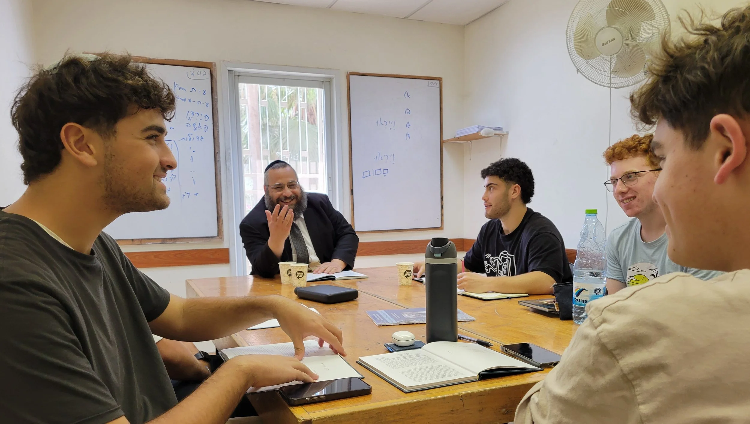 A group of young men and a middle-aged man sitting around a table in a room, engaging in conversation and smiling. The room has whiteboards and a window with bars, a fan on the wall, and various items like notebooks, books, and bottles on the table.
