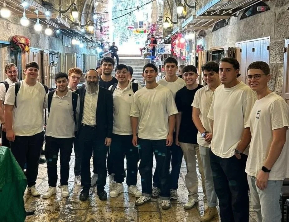 Group of young men and an adult man standing in a decorated indoor market or corridor, some wearing white shirts, with string lights and colorful decorations overhead.