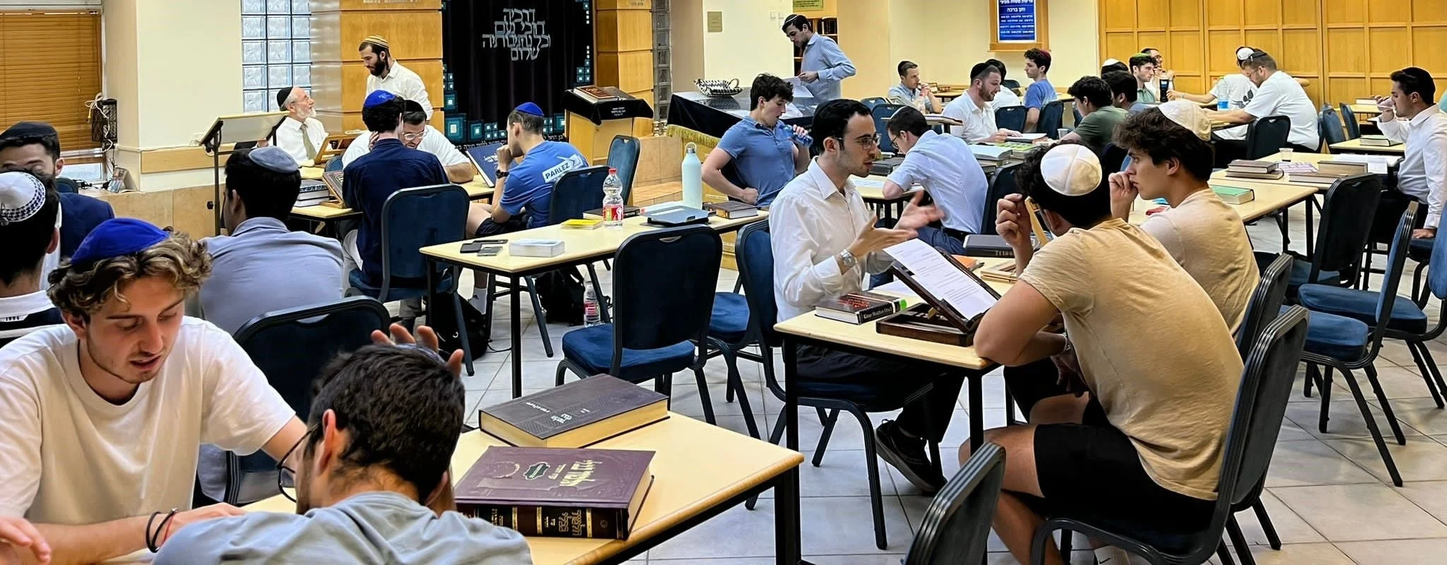 Young Jewish men studying and discussing in a spacious room with many tables, books, and some wearing kippahs.