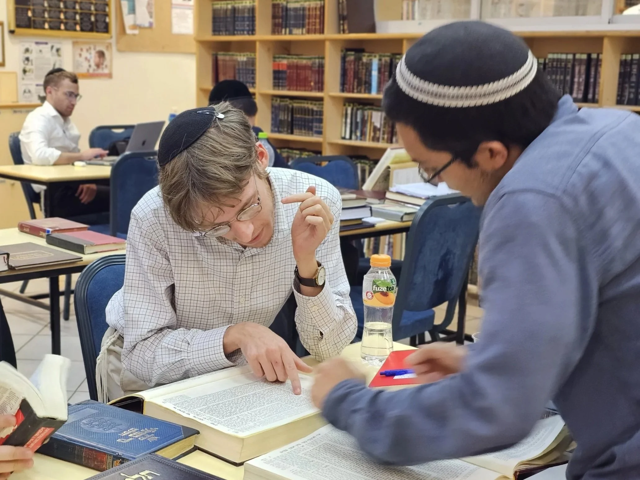 A diverse group of people, including a young man with glasses and an older woman wearing a kippah, gathered around a table in a library or classroom, engaged in study and discussion with open books, notebooks, and a water bottle present.