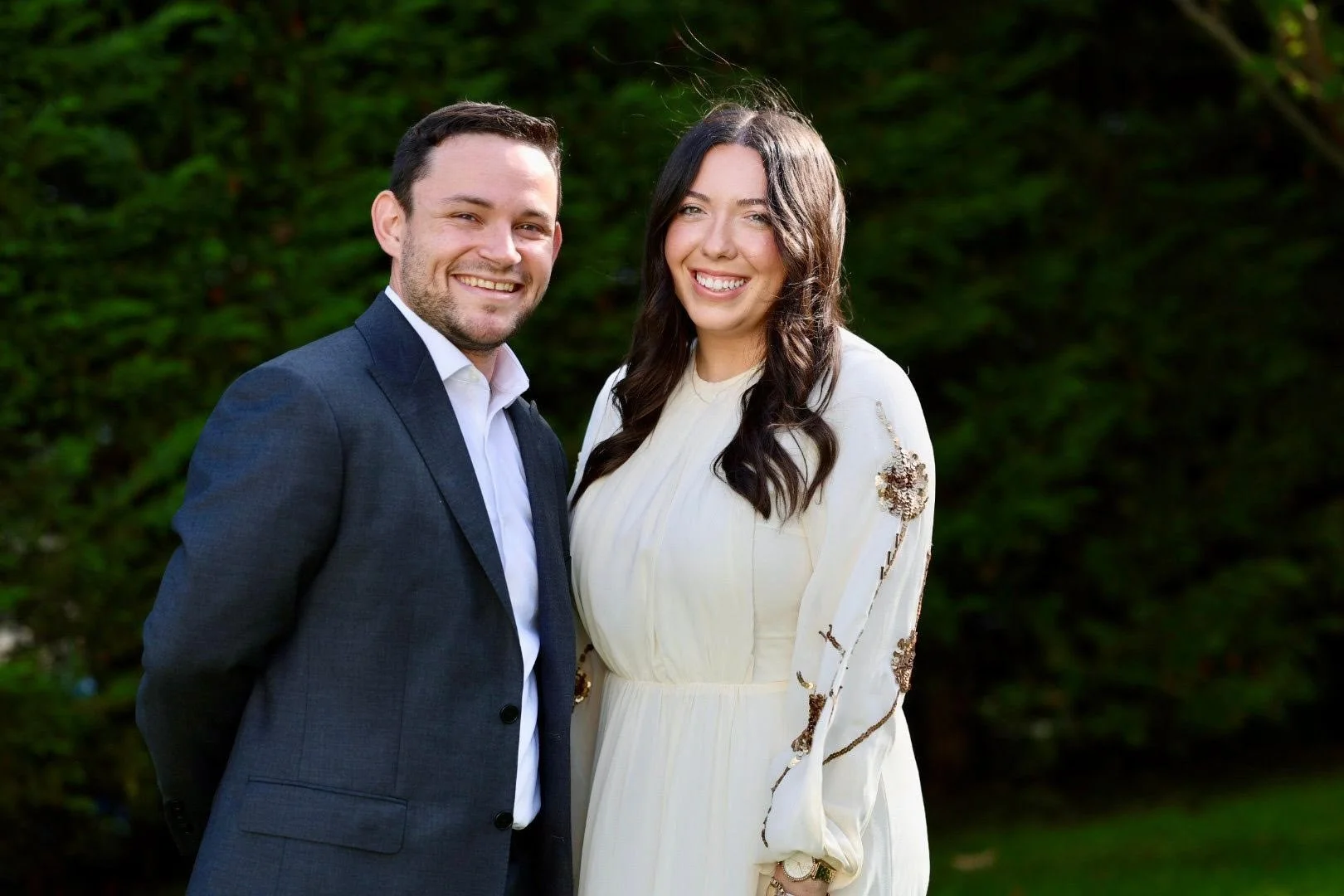 A smiling man and woman standing outdoors with a green leafy background.