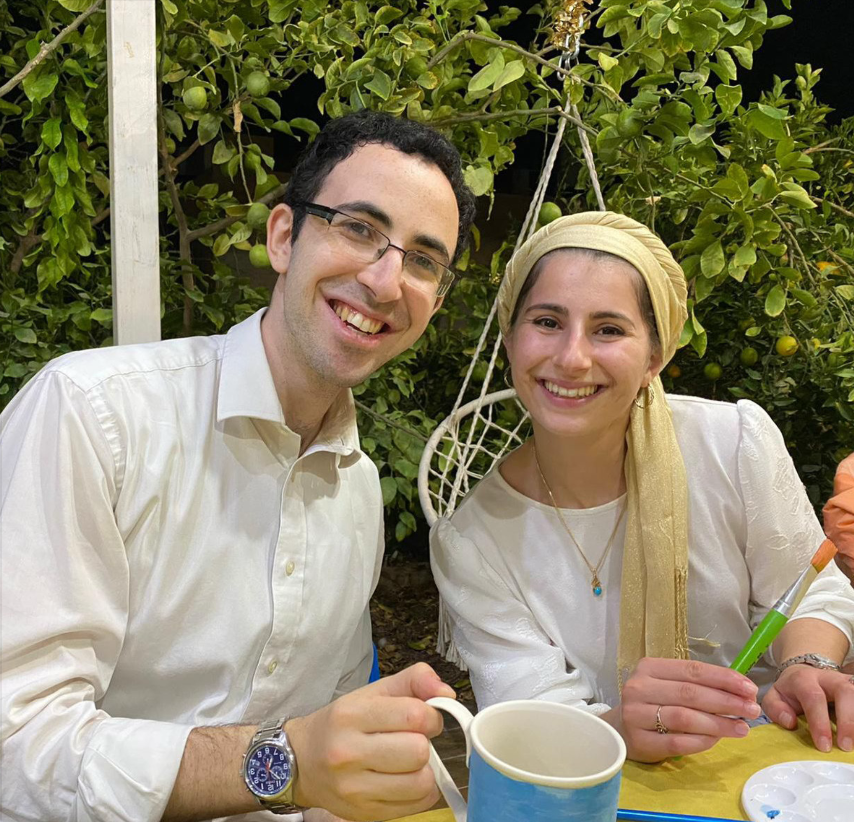 Two individuals sitting at a table outdoors, smiling at the camera, with green foliage and fruit trees in the background, appear to be enjoying a social gathering.
