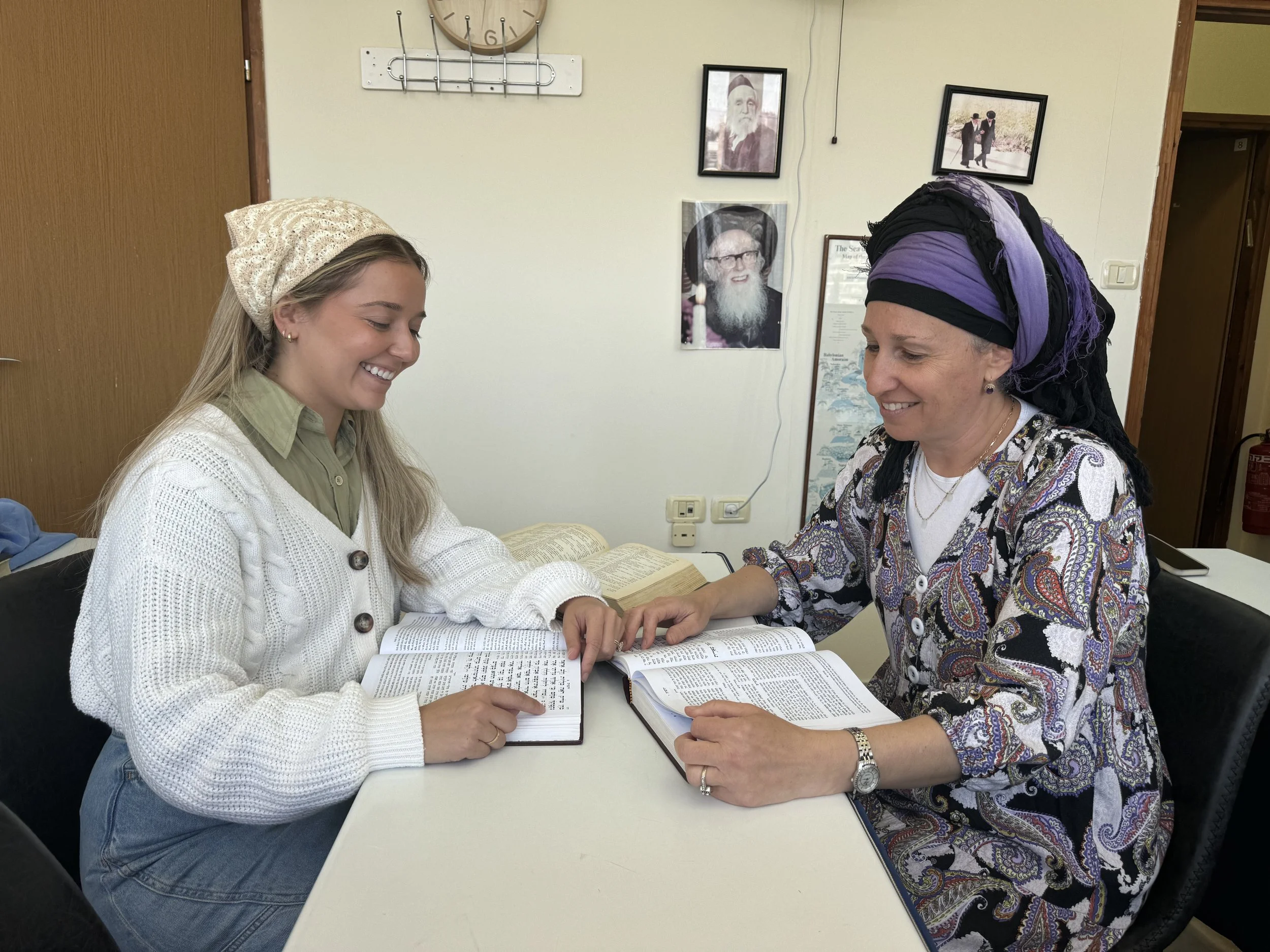 Two women smiling and looking at open books on a table. One woman is wearing a light-colored headscarf and a green shirt, and the other is wearing a black and purple headscarf with a patterned blouse. They are indoors, with framed black-and-white photographs on the wall behind them.