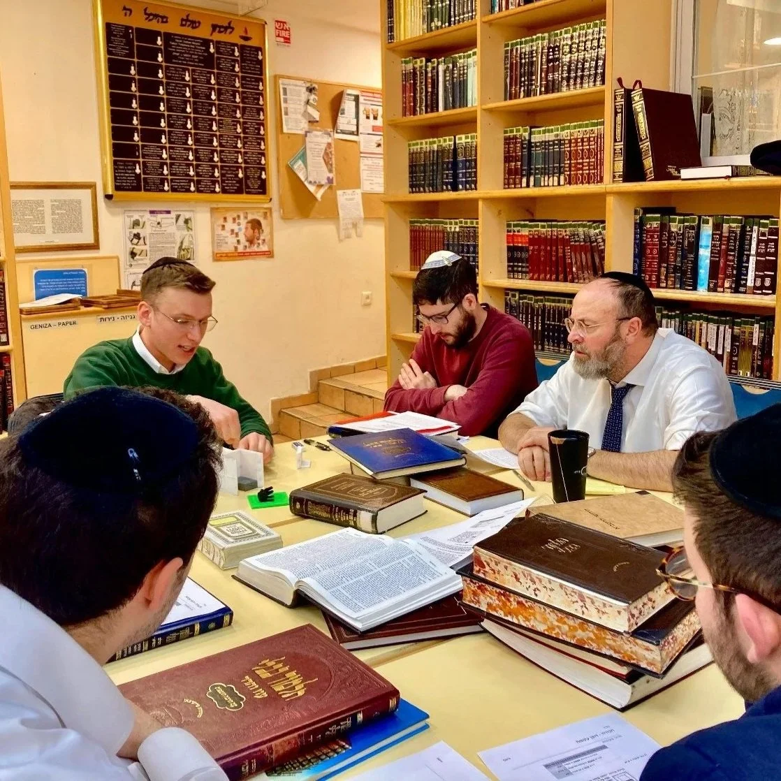 Group of men with religious attire and books, sitting at a table in a library or study room, engaged in discussion or learning.