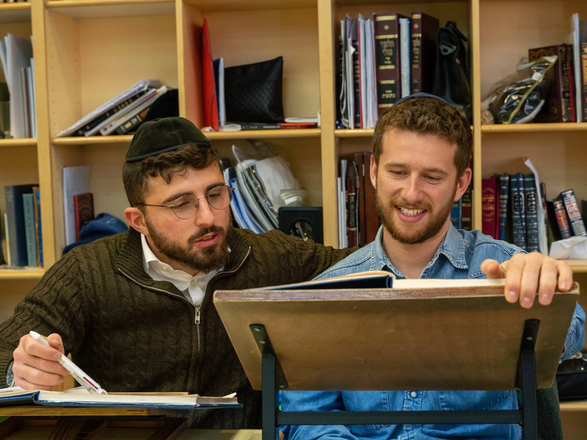 Two young men sitting at a table behind a wooden bookshelf filled with books and folders, engaging with a tablet or electronic device.