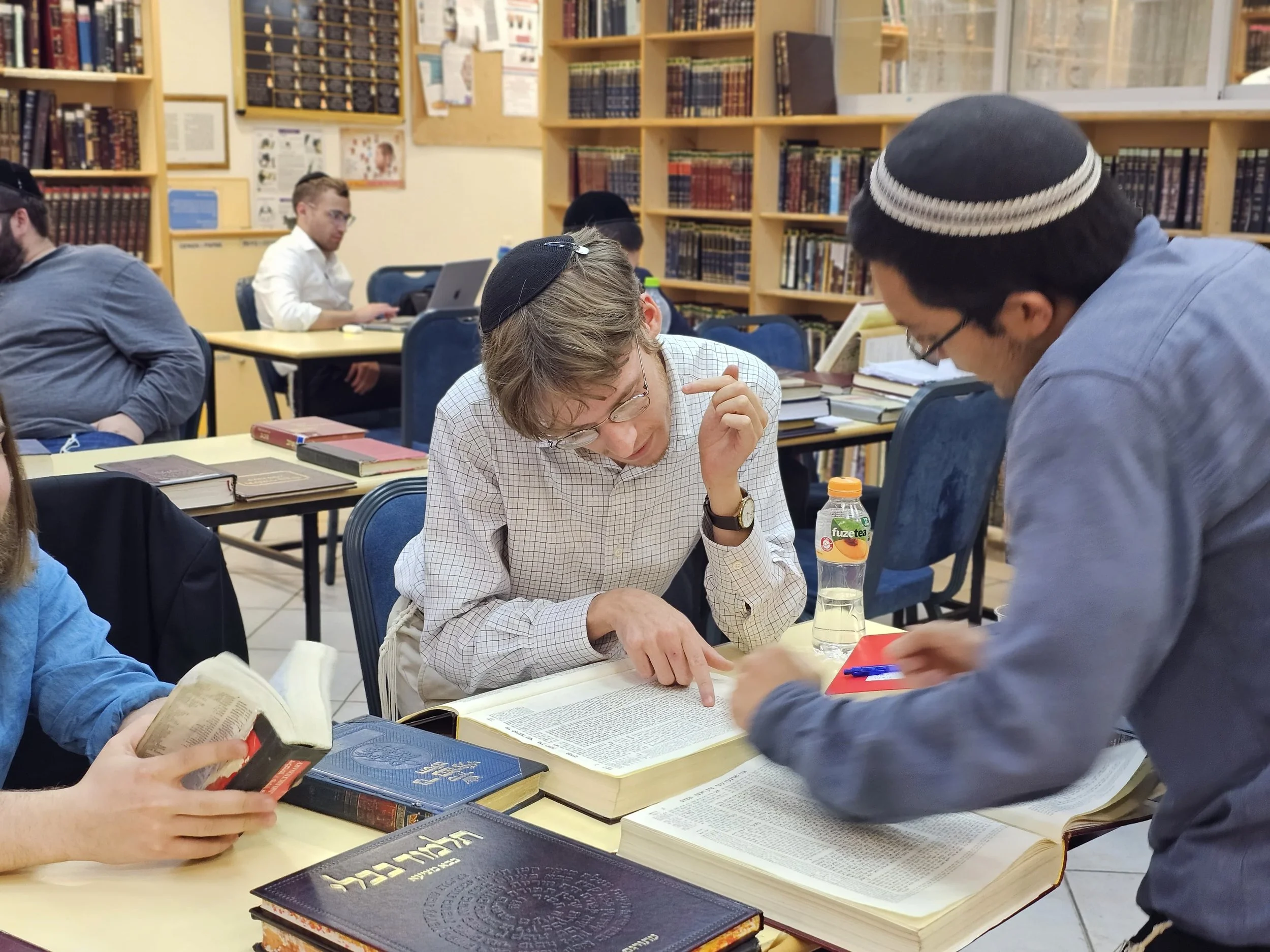 Several individuals wearing kippahs are seated at tables in a library, engaging with books and talking. Shelves of books are visible in the background.