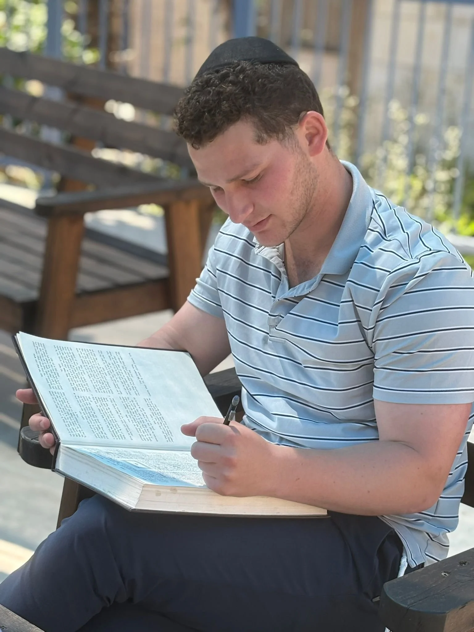 A young man with curly dark hair wearing a black kippah and a striped polo shirt, sitting outdoors on a bench, looking down at a book he is reading.