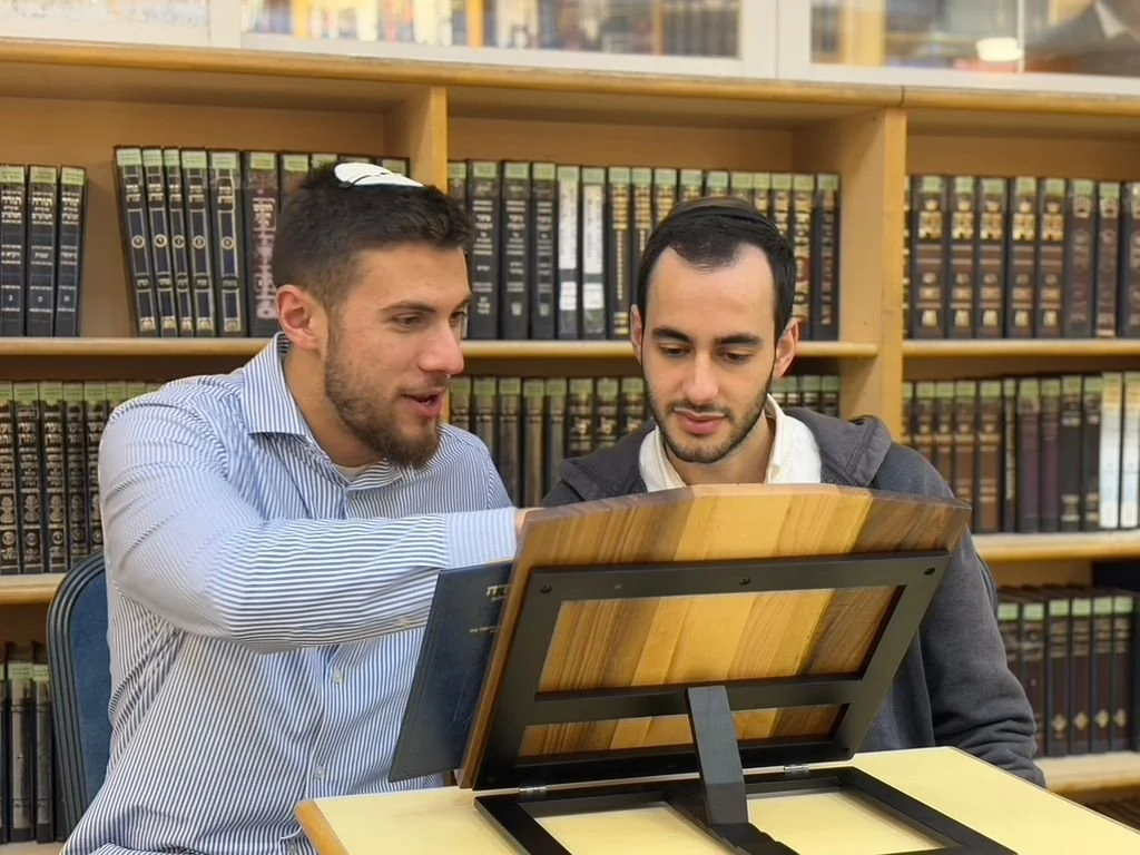 Two men are sitting at a table, looking at an artwork or a display. They are in a library or bookstore with bookshelves filled with books in the background.