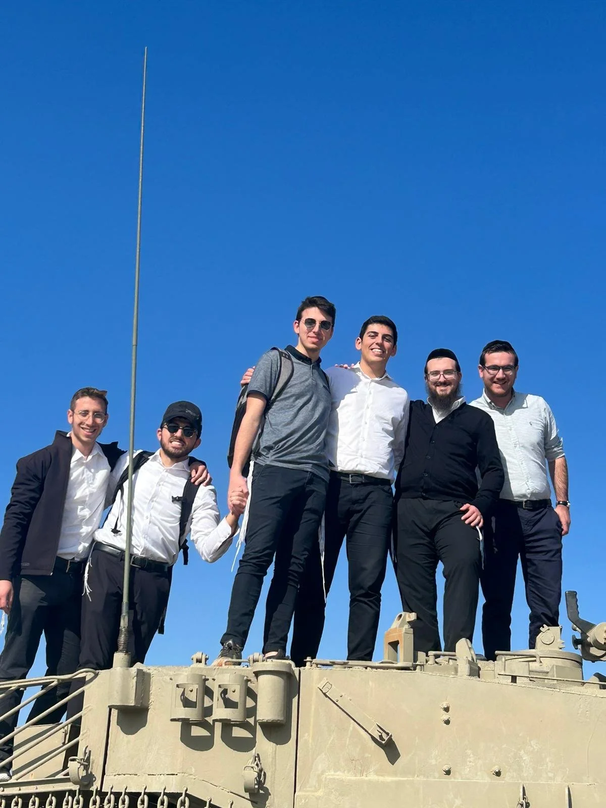 Six young men standing together on top of a military tank, smiling, dressed in smart-casual attire, with a clear blue sky in the background.