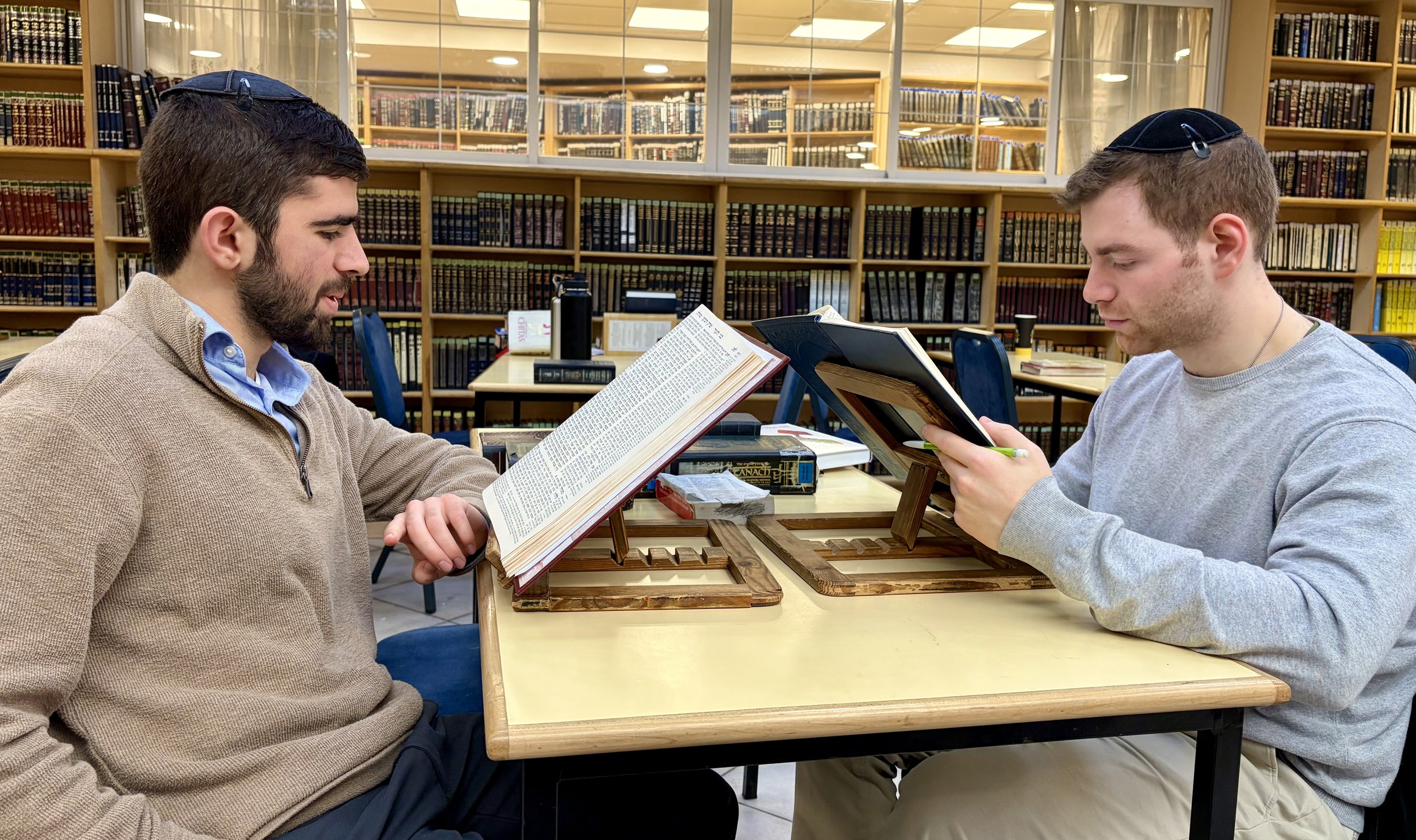 Two men wearing kippahs study religious texts at a table in a library, with shelves of books in the background.