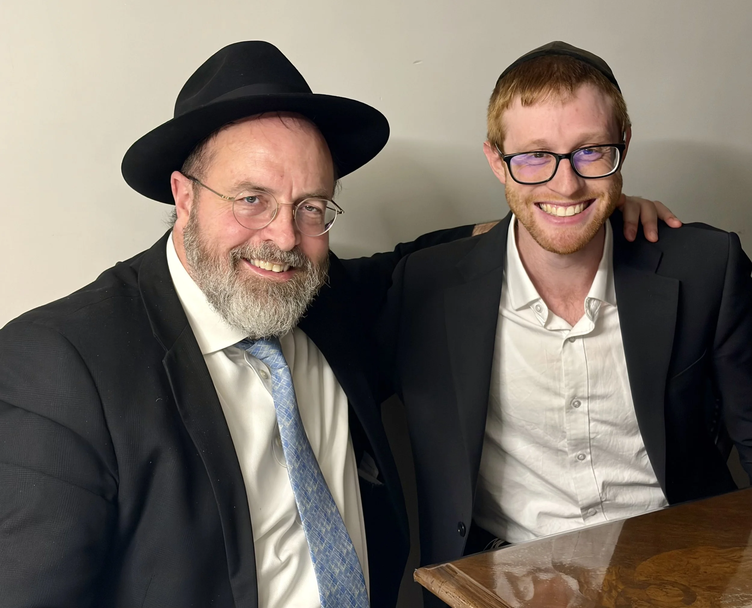 Two men wearing black suits, white shirts, and glasses, with one man having a beard and the other with short red hair and a beard, both smiling and posing together. The man on the left is wearing a black hat, and the man on the right has his arm around the other man's shoulder.