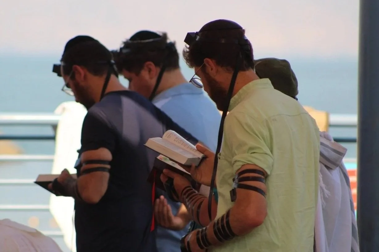 Three men with tefillin on reading prayer books near a railing, with a body of water in the background.