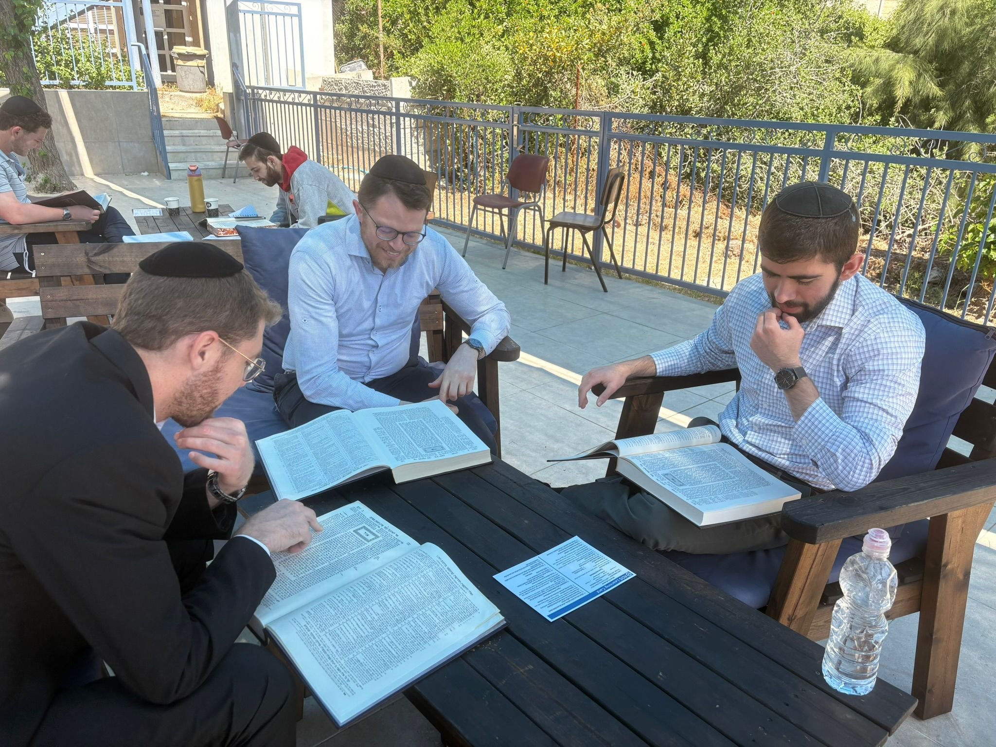 A group of men with yarmulkes reading religious texts outdoors at a wooden picnic table, with trees and bushes in the background.